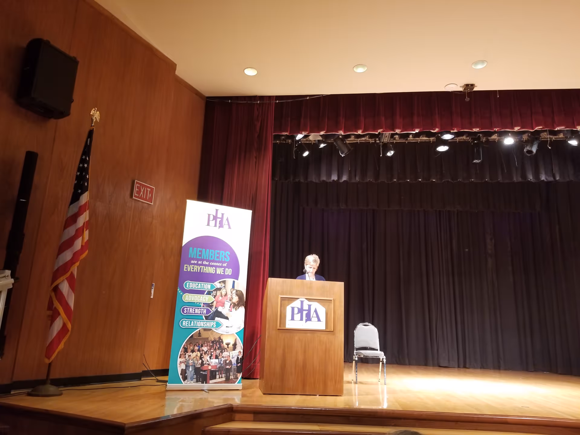 A speaker stands at a podium on a stage with curtains, banner, and American flag.