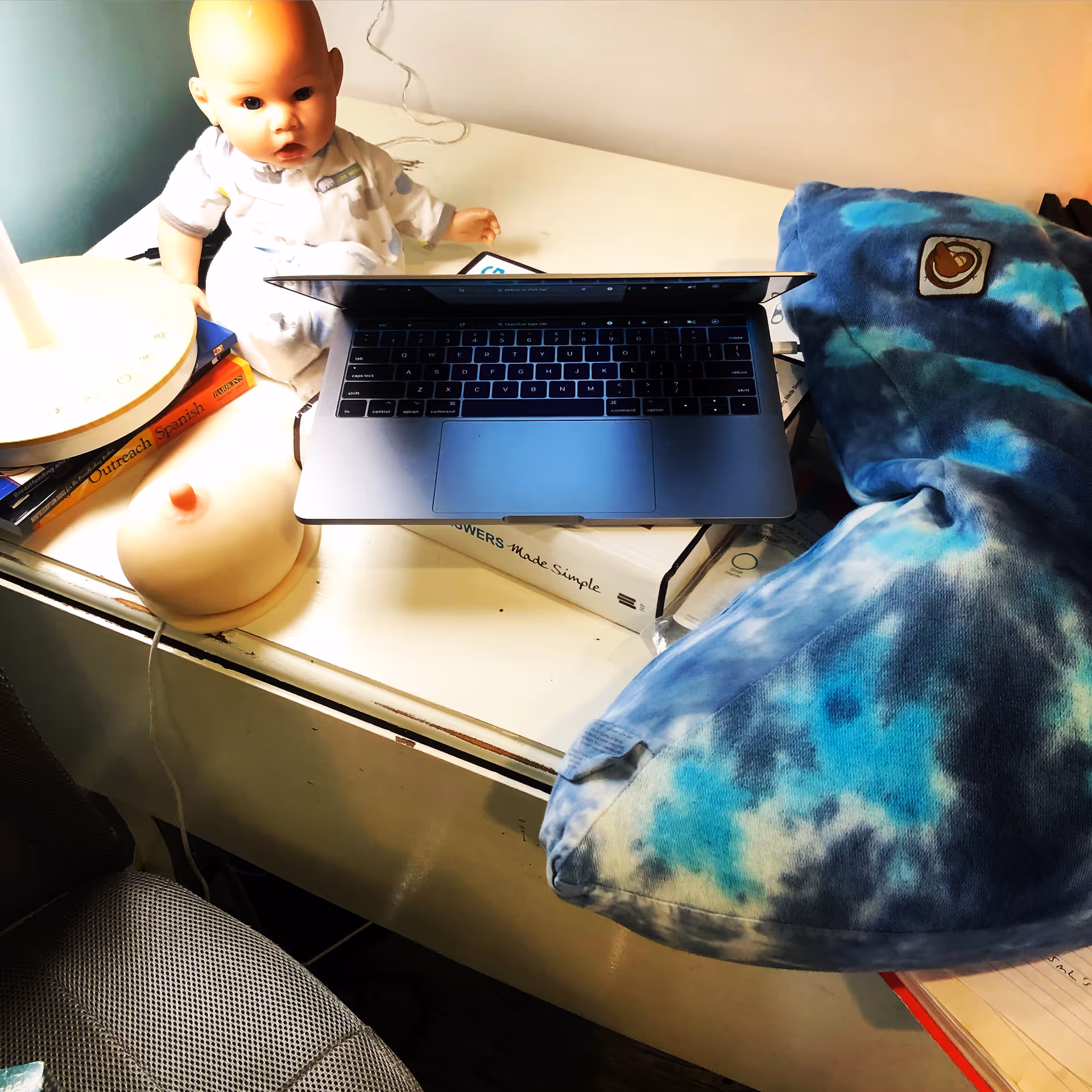 A laptop rests on a cluttered desk beside a baby doll and a blue tie-dye blanket.