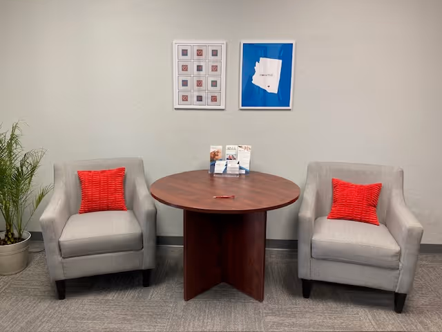 Two gray armchairs with red cushions flank a round wooden table in a neutral waiting area.