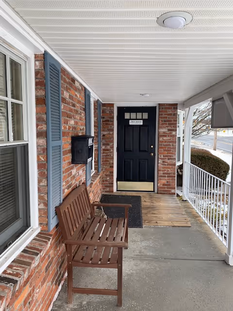 Brick front porch of a facility with a wooden bench, mailbox, and dark front door.
