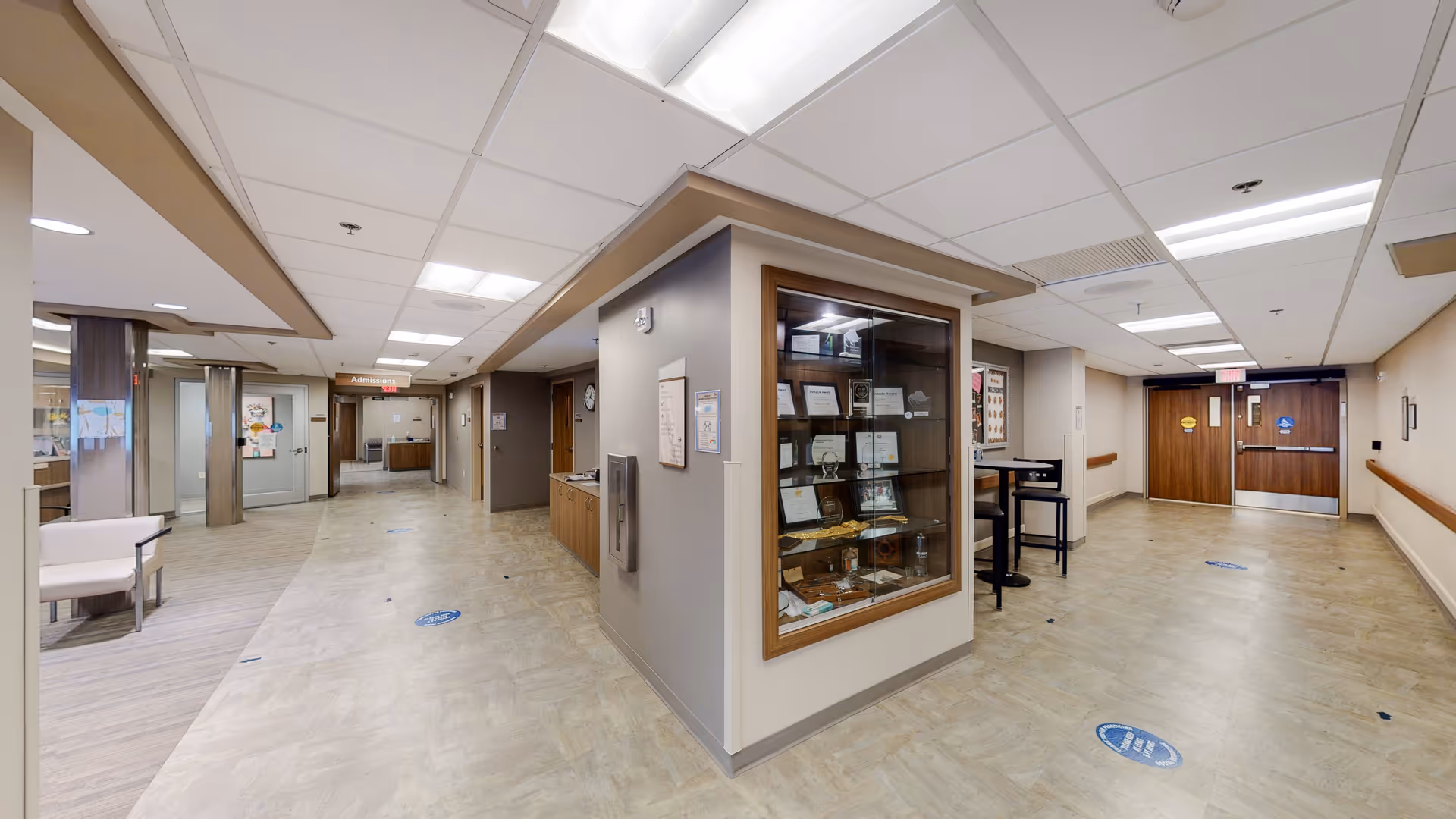 Wide interior corridor of a hospital-like facility with display cabinet and distant double doors.