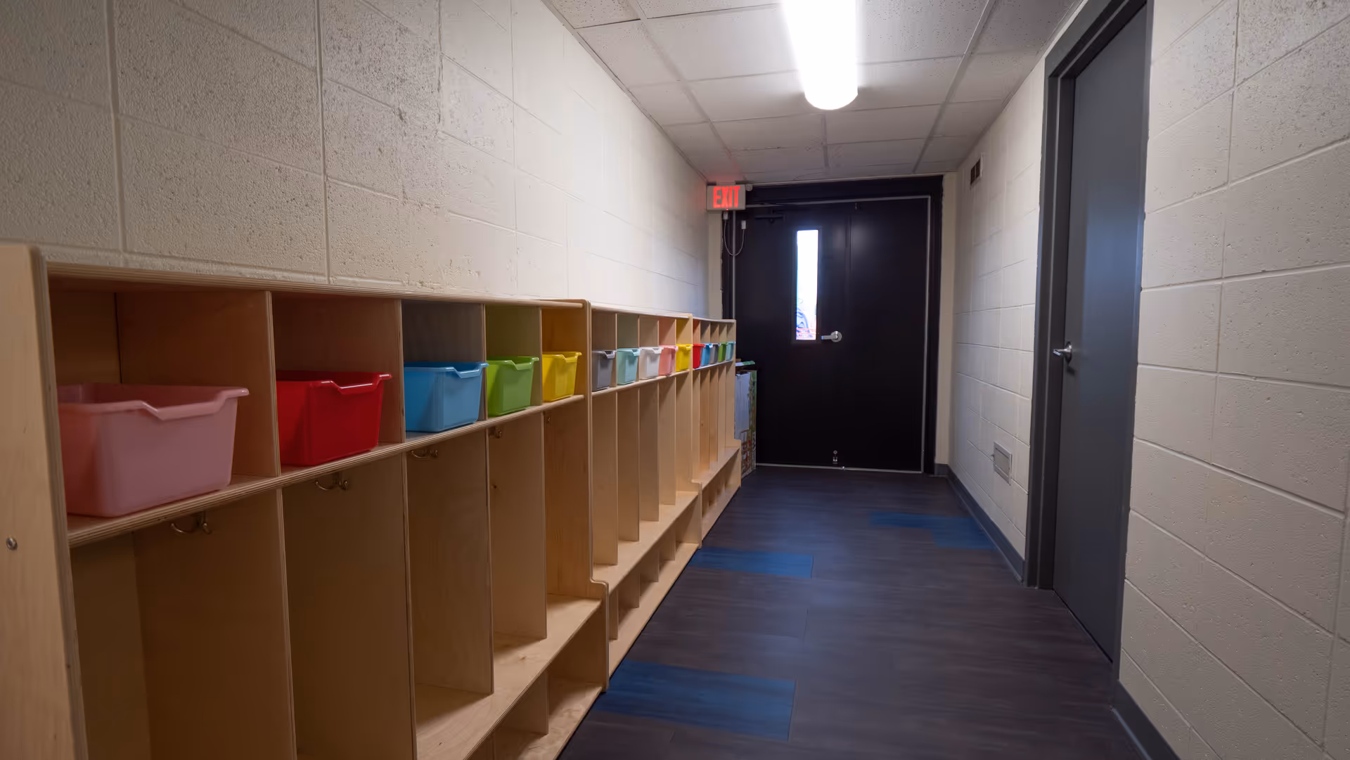 Long hallway with wooden cubbies and colorful plastic bins along the left wall, door at end.