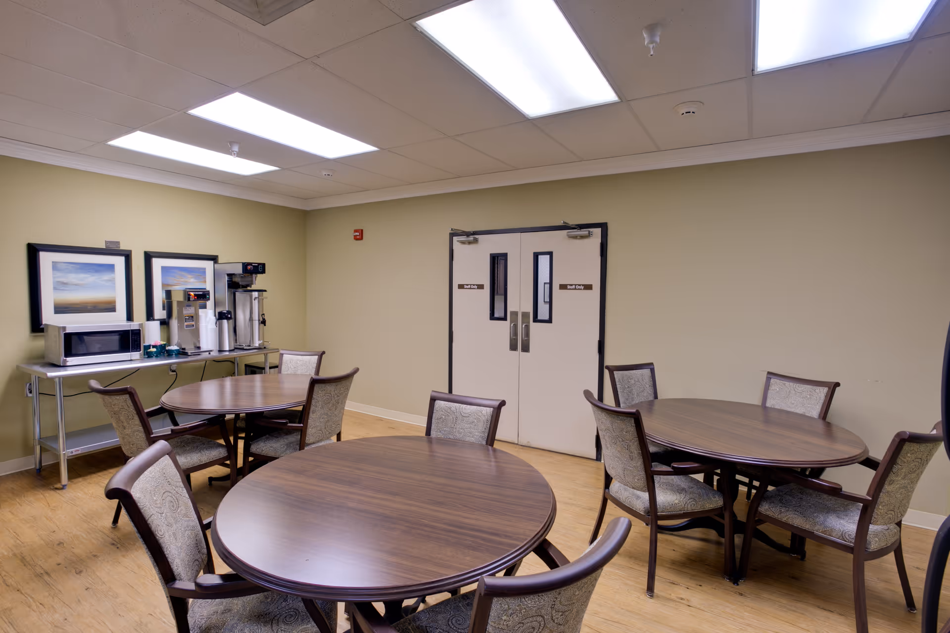 A dining area with round wooden tables, chairs, and a beverage station along the wall.