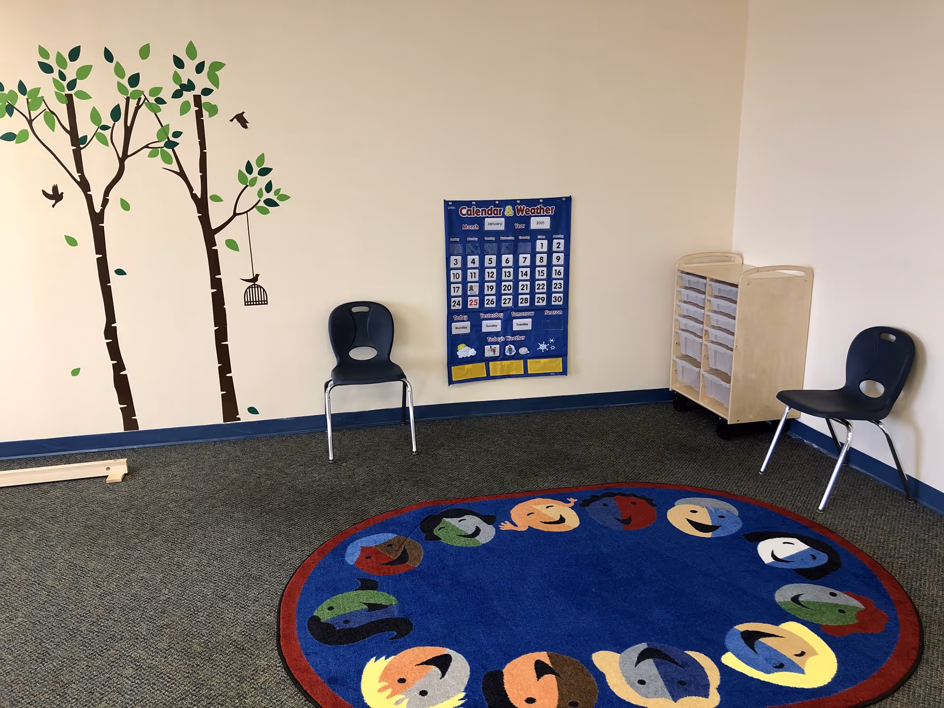Interior play area with wall mural, calendar, chairs, and colorful circular rug.