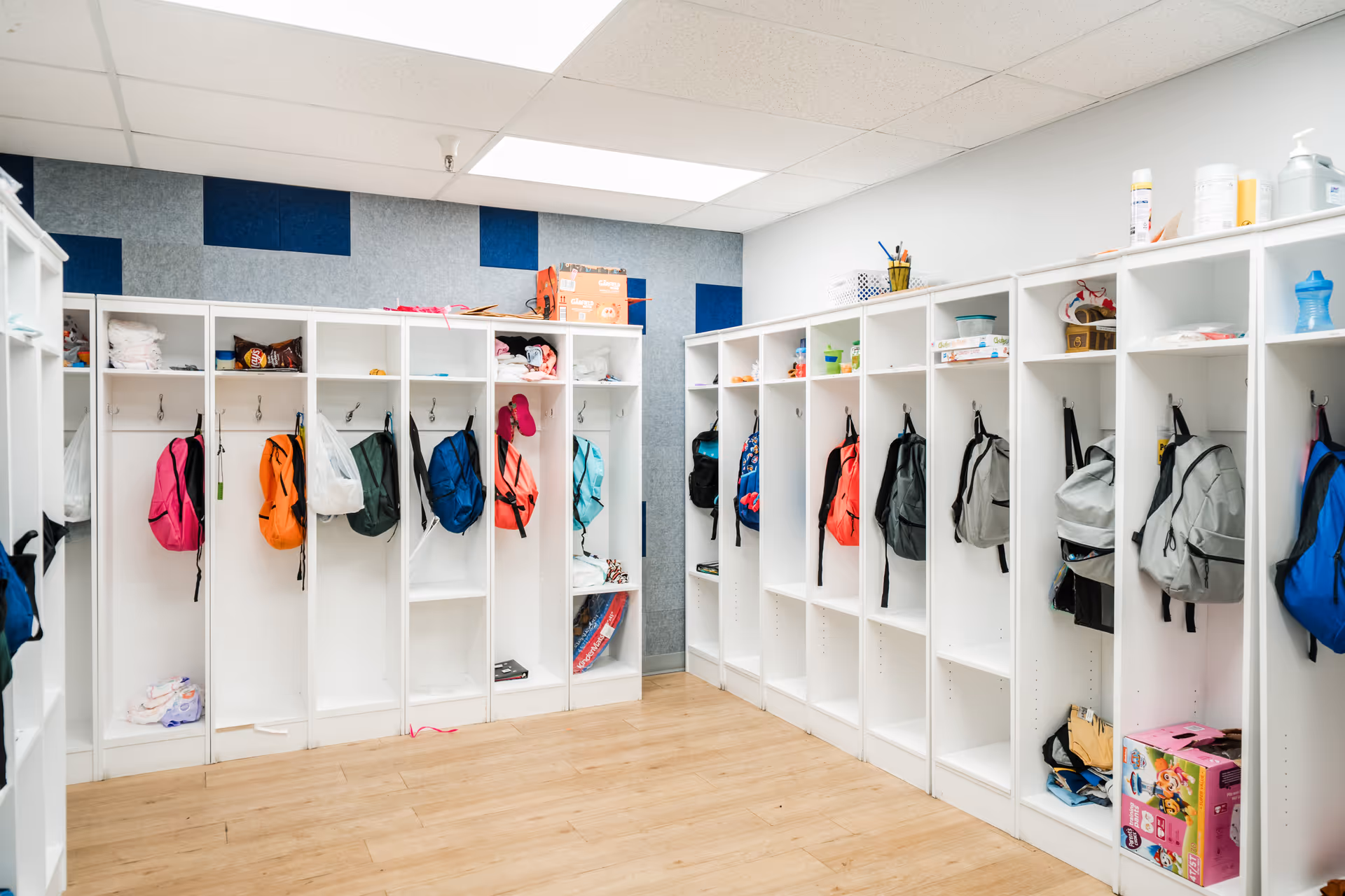 Row of white cubby lockers with hanging backpacks in a bright facility hallway.