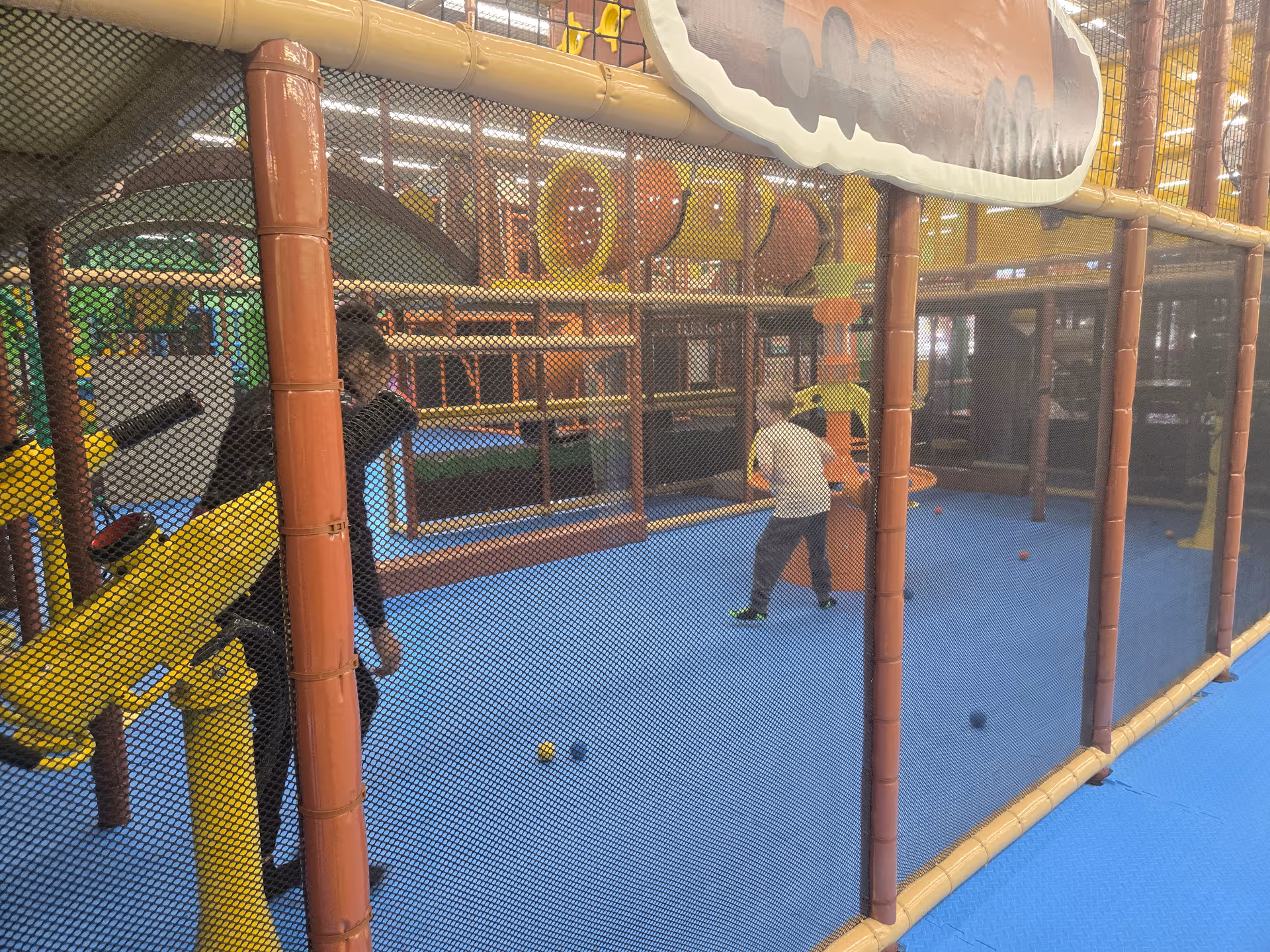 Child and adult supervise a padded indoor playground enclosed by netting.