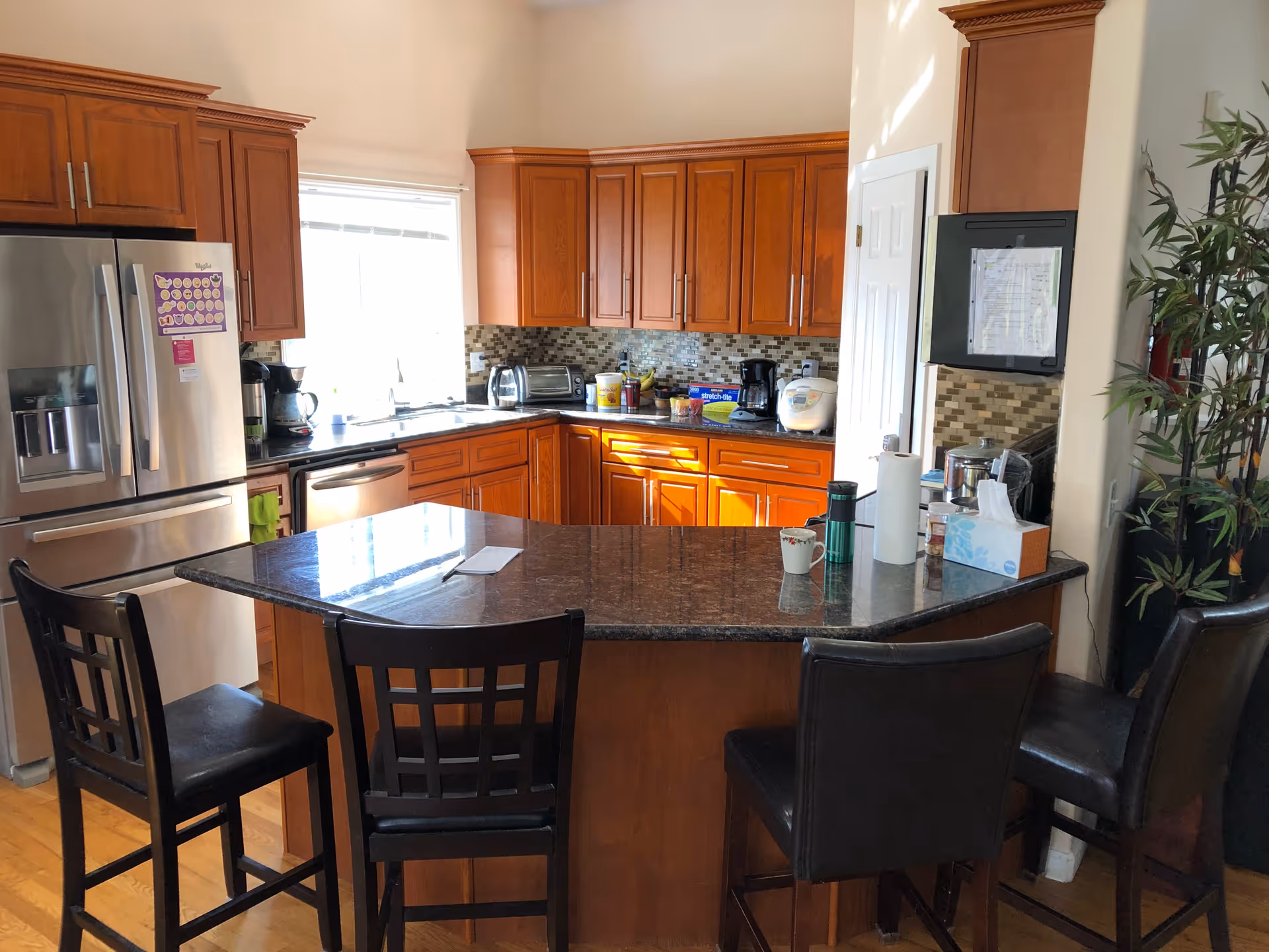 Open kitchen with wooden cabinets, granite island, stainless appliances, and bar stools.