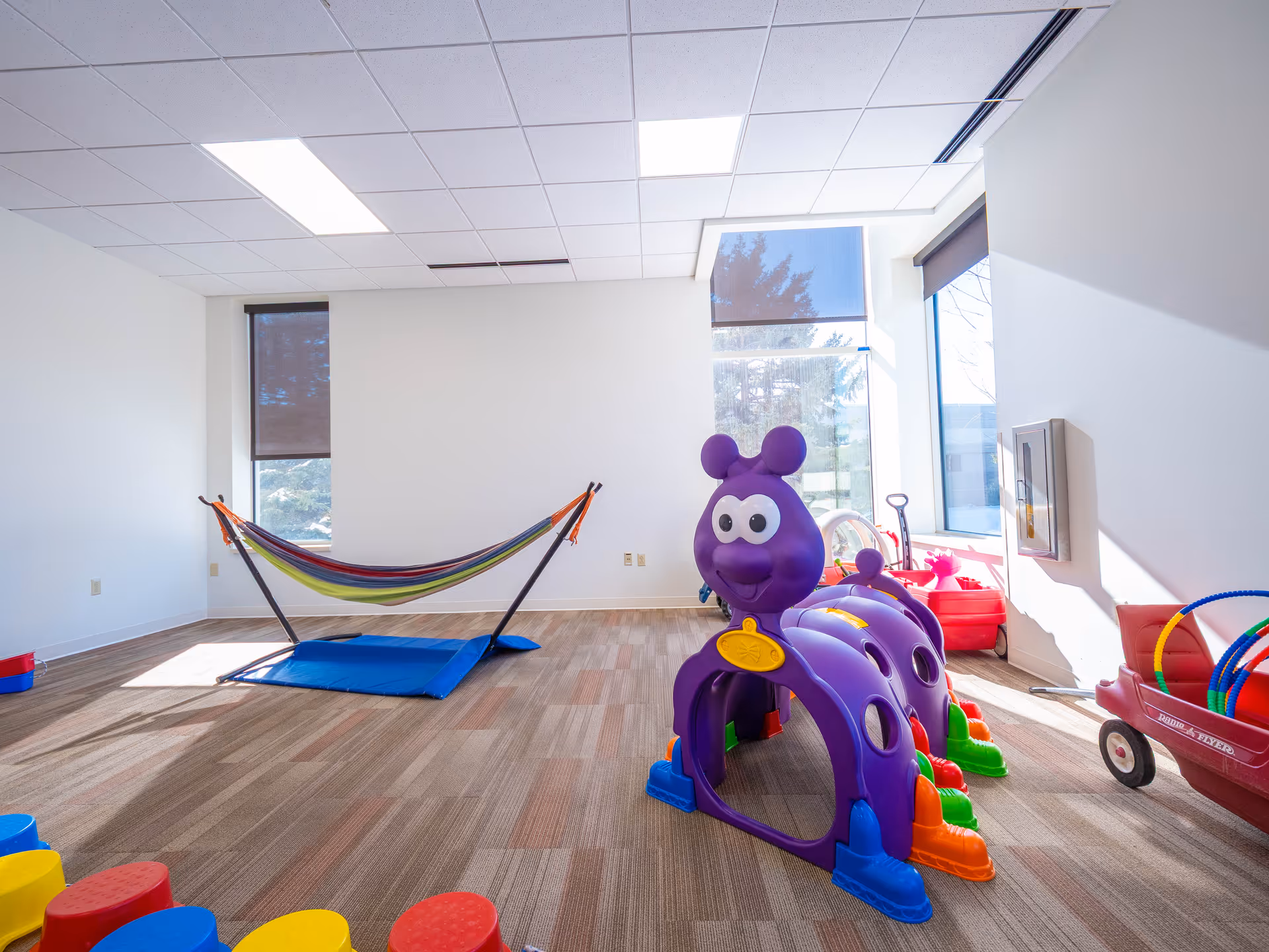 Bright indoor playroom with colorful toddler toys, a hammock, and large windows.
