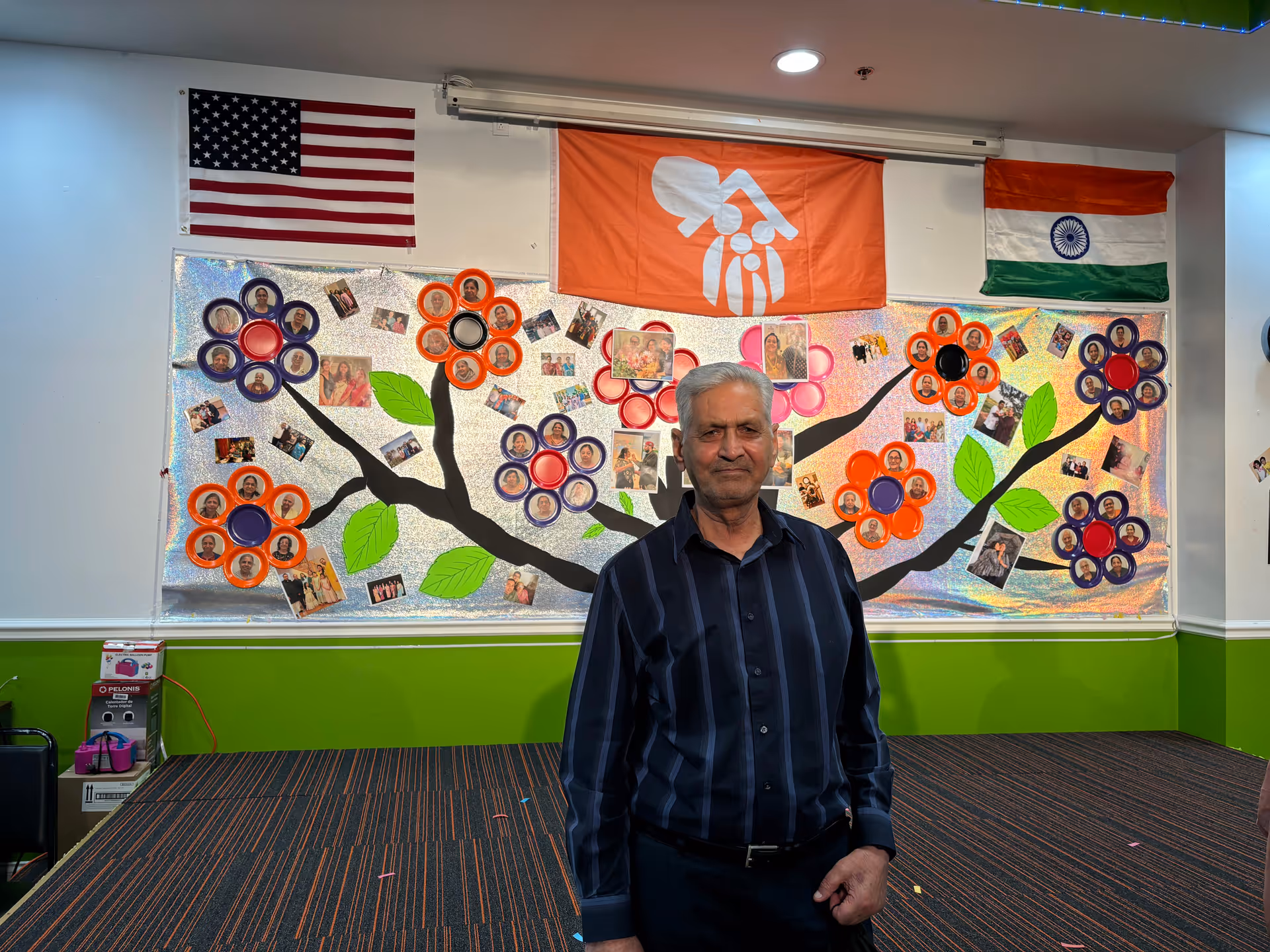 A man stands in a decorated interior with a mural, flags, and photos on the wall.
