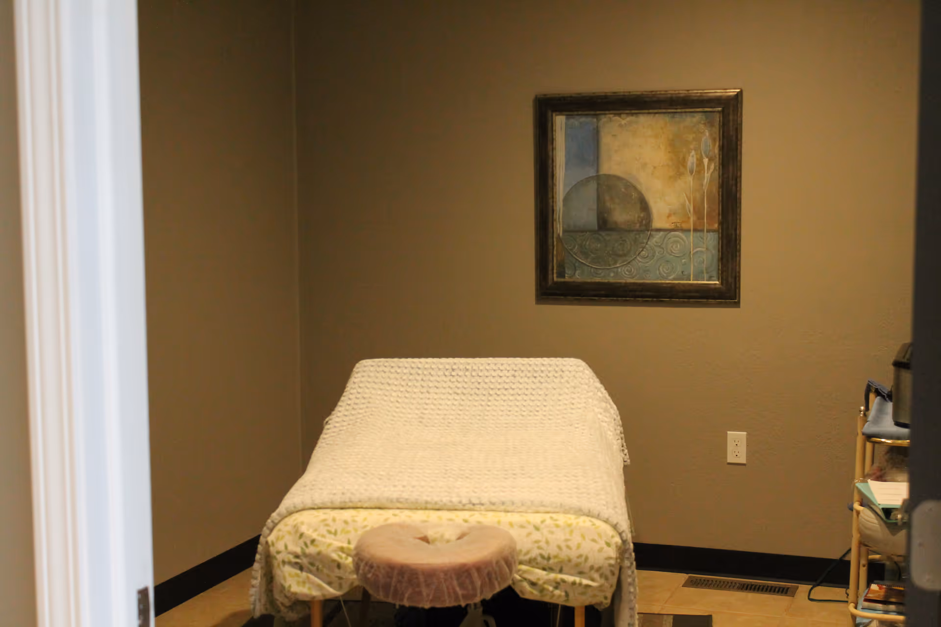 A treatment room features a bed-like table covered with a blanket and framed wall art.