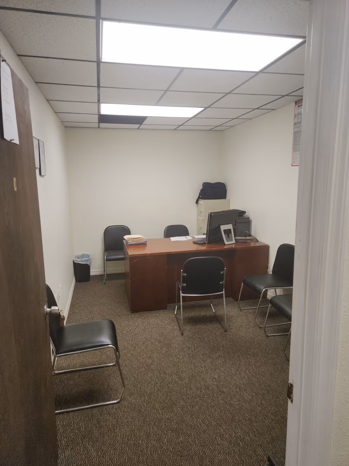 Small office with a desk, computer, and several black chairs in a waiting area.