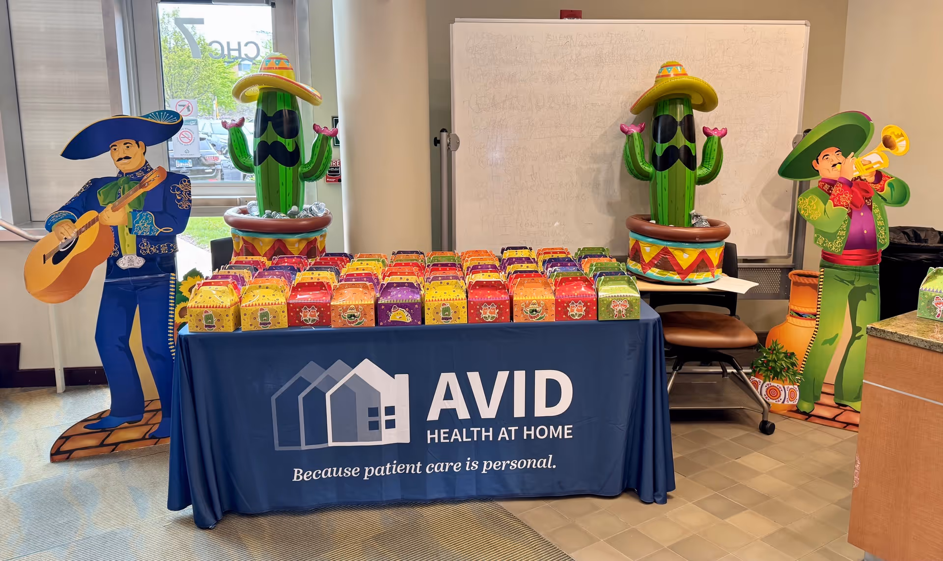 Indoor table setup featuring AVID Health at Home banner, colorful gift boxes, and cactus decorations.