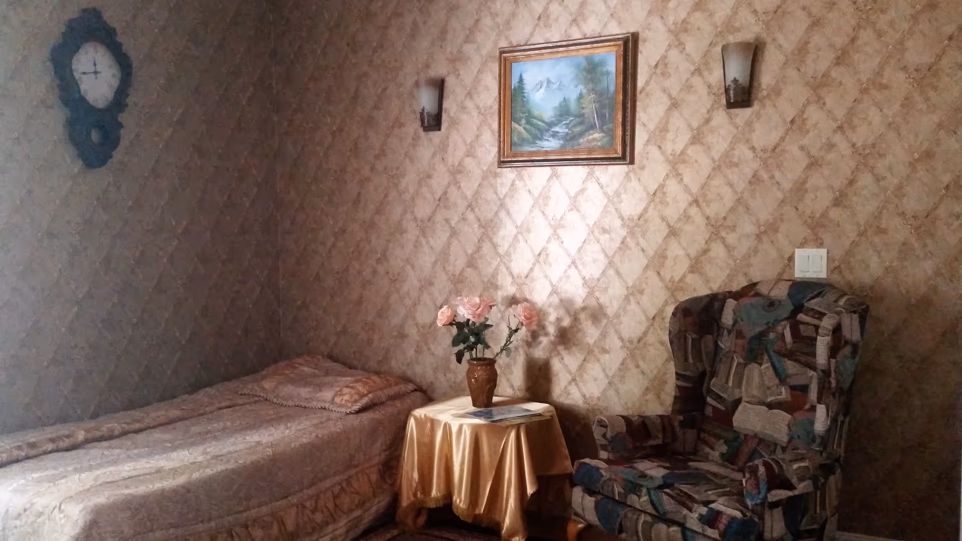 A small bedroom with a bed, armchair, round table with pink flowers, and floral wallpaper.