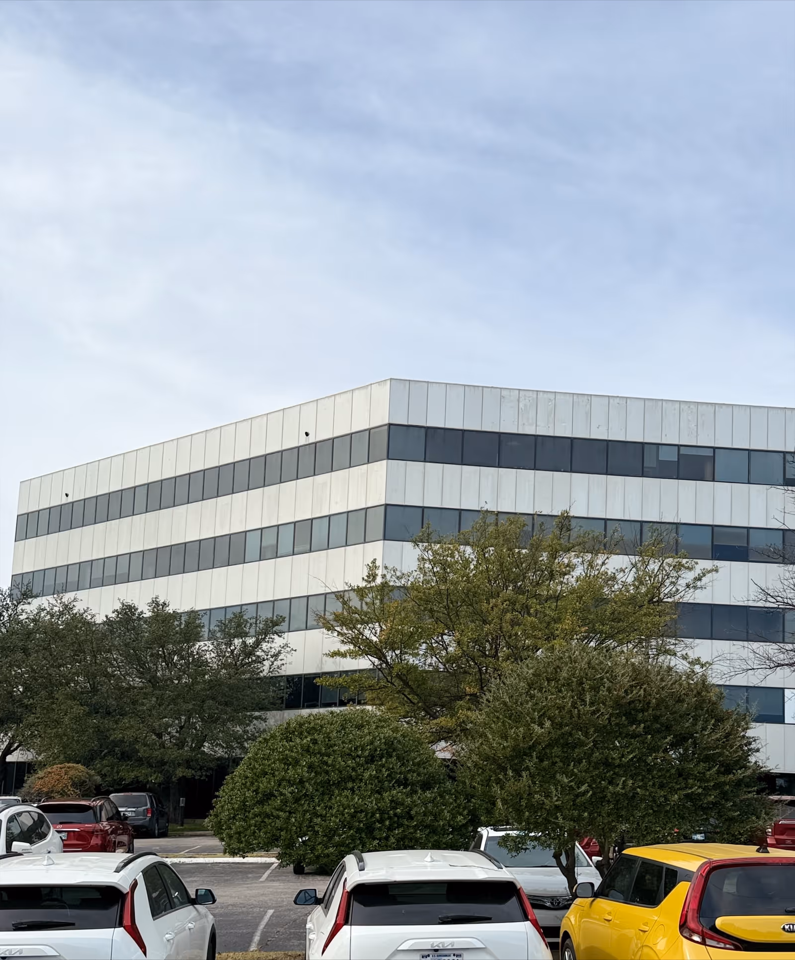 Modern multi-story office building with glass windows, trees, and parked cars in foreground.