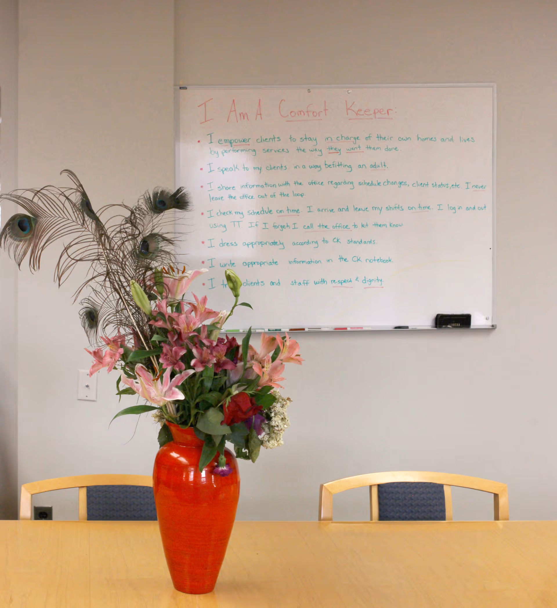 Bright interior with a wooden table, red vase, flowers, and peacock feathers.