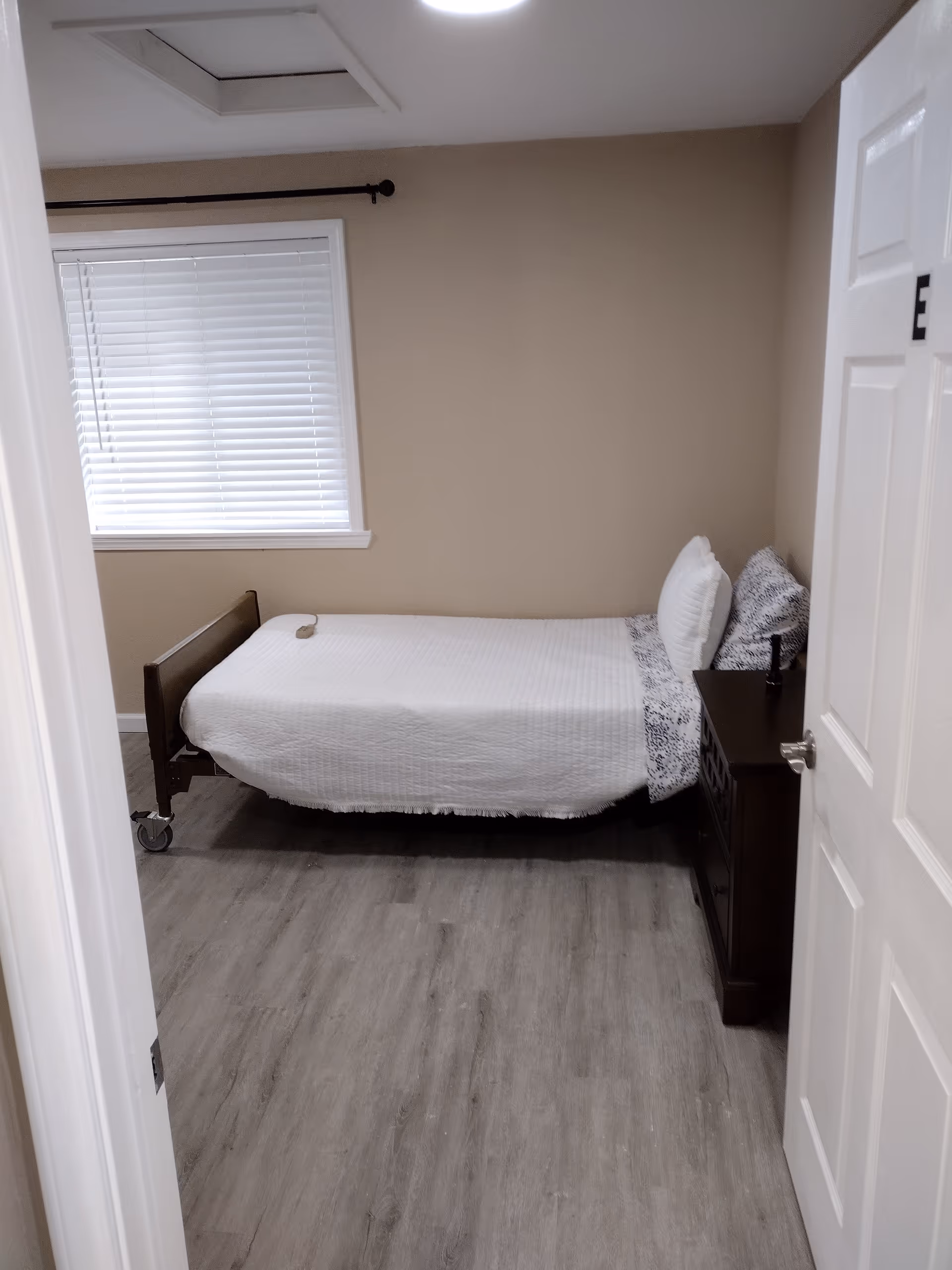 Small bedroom with single bed, nightstand, window blinds, beige walls in care facility