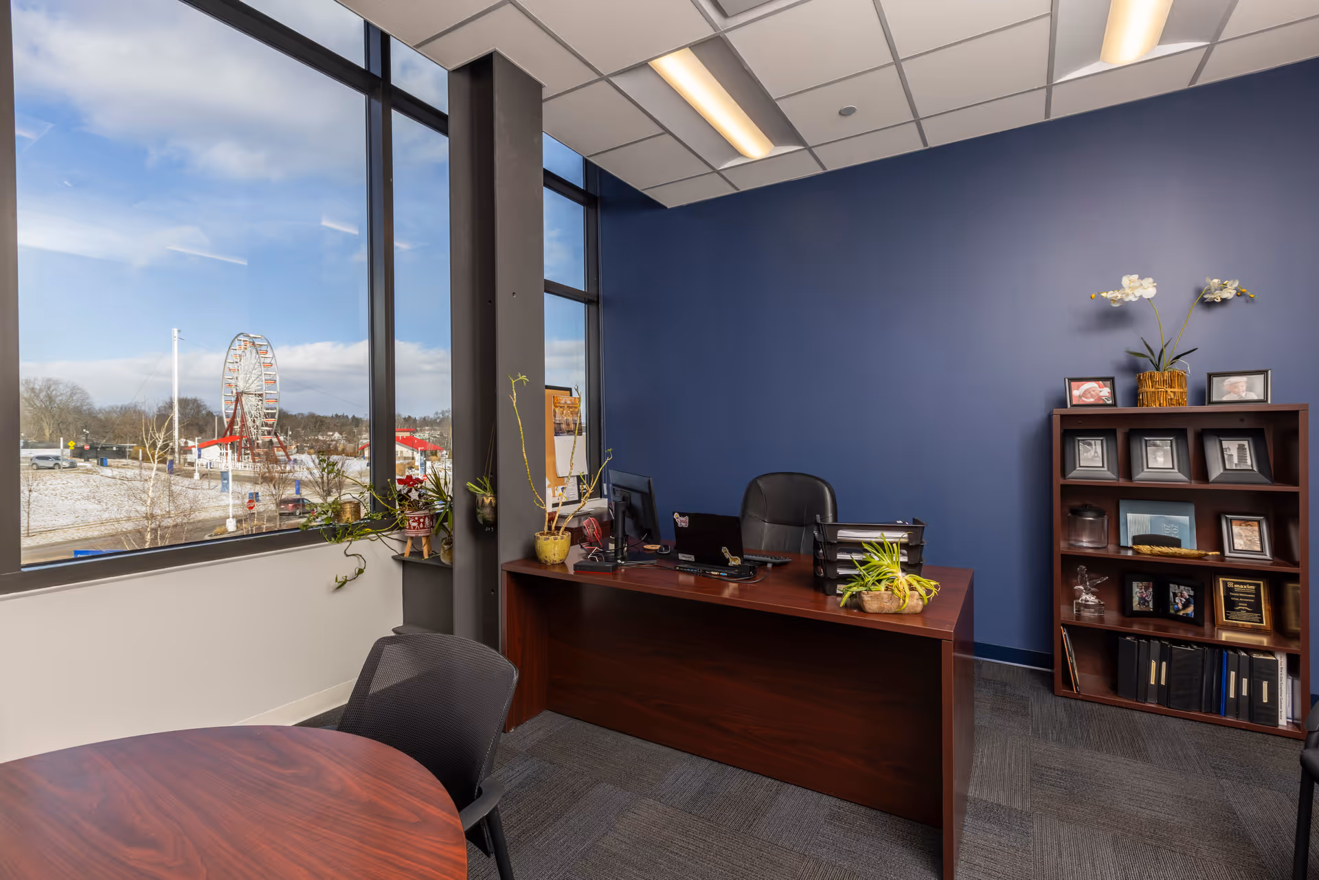 Indoor office reception with desk, round table, plant, and large windows overlooking outdoors.