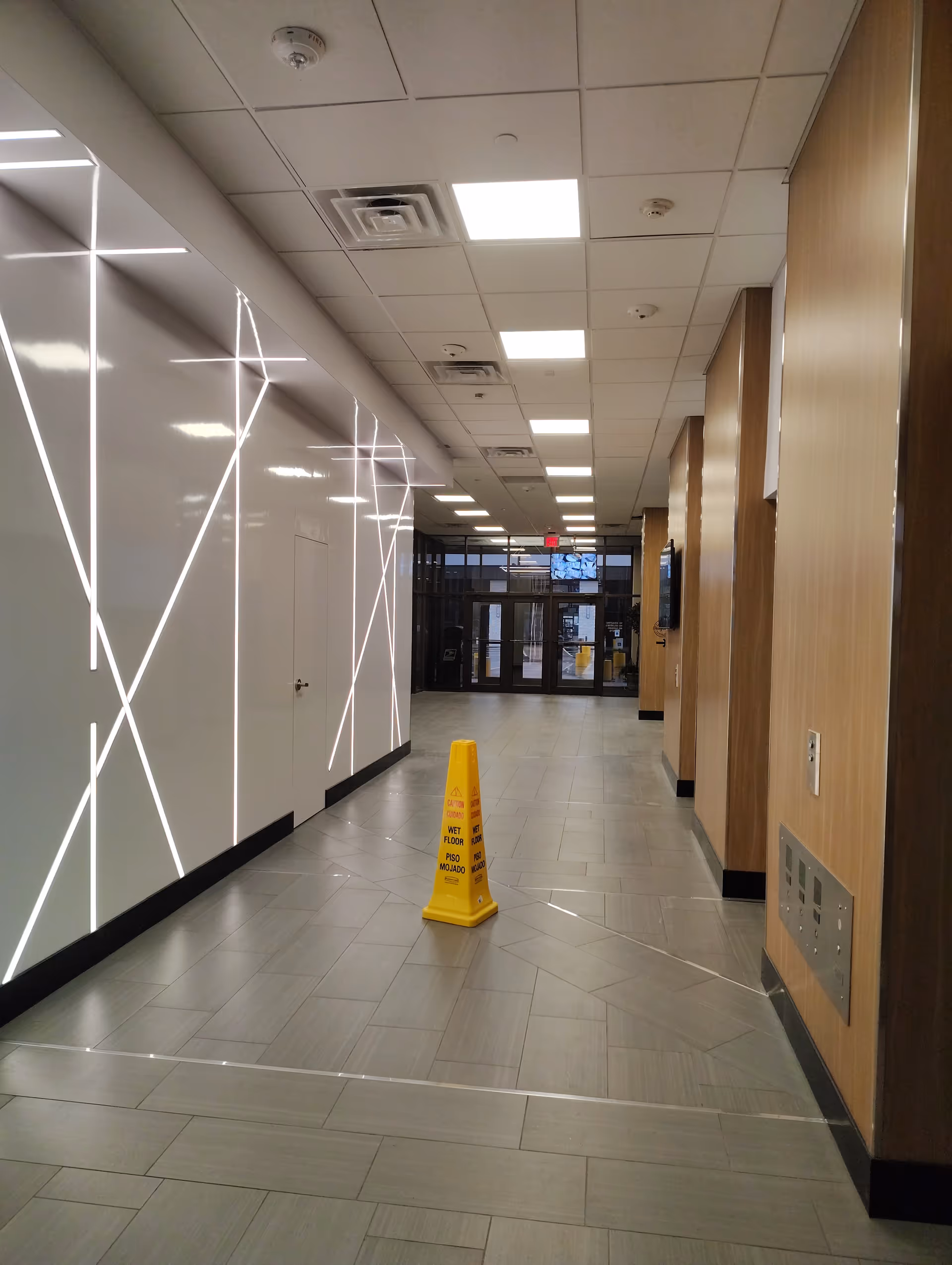 Indoor hallway with illuminated diagonal wall lines, wood-paneled walls, and a yellow wet floor sign.