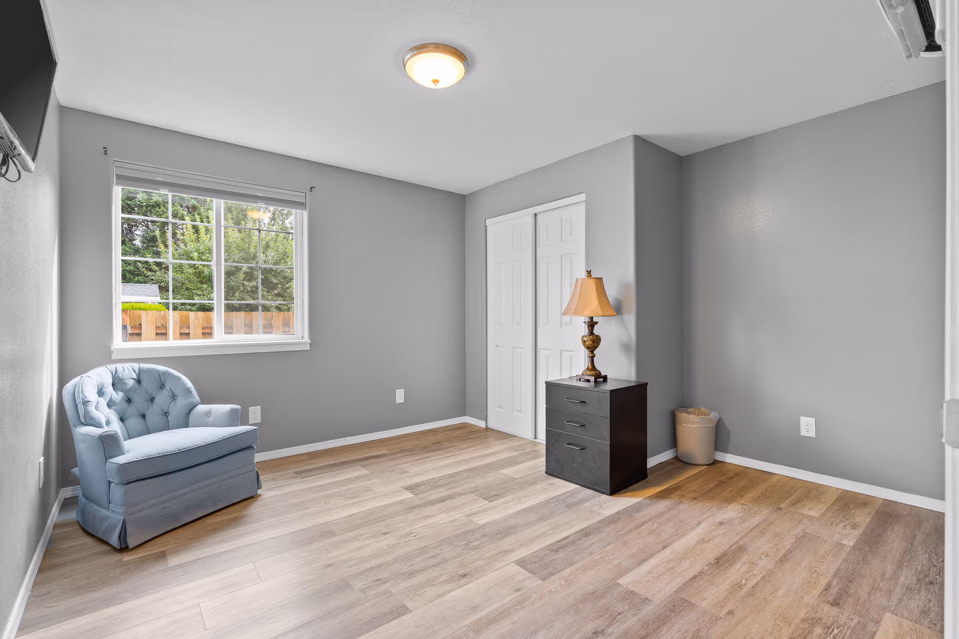 A light gray bedroom with a blue upholstered armchair, dresser, and a window.