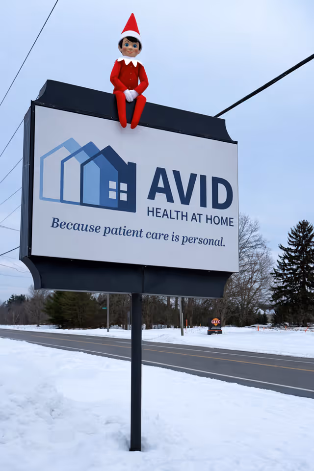 Outdoor sign for AVID Health at Home with a red elf figure on top in snow
