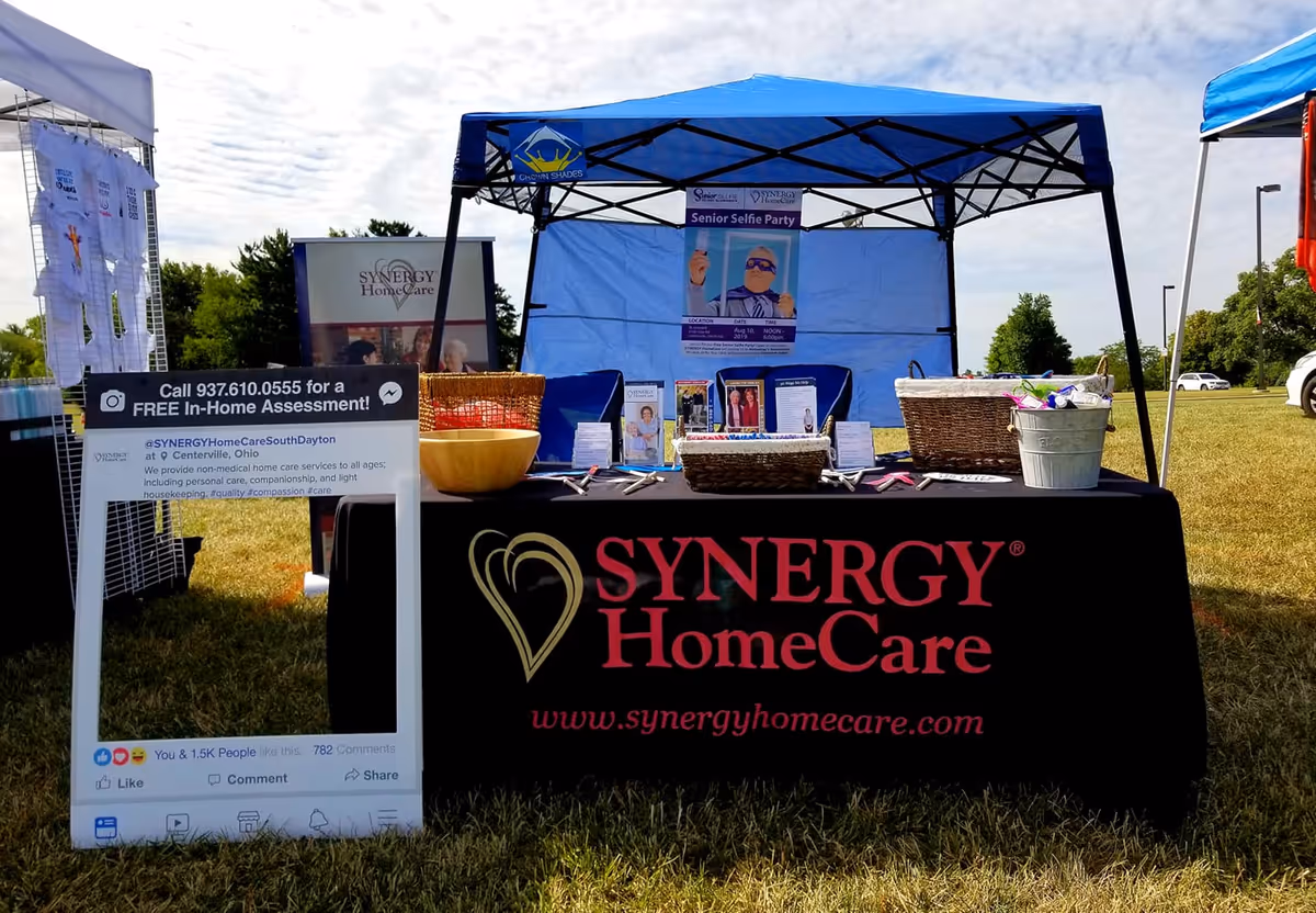 Outdoor SYNERGY HomeCare booth with a black branded table under a blue canopy.