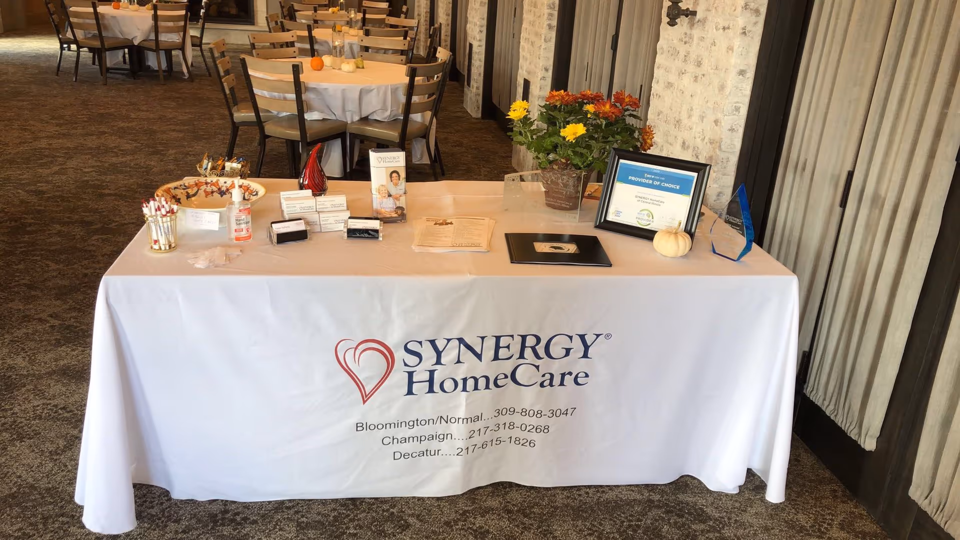 Indoor event setup with a Synergy HomeCare table, materials, and chairs in the background.