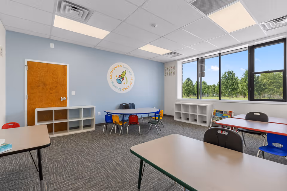 Bright classroom-style activity room with tables, colorful chairs, and large windows overlooking trees