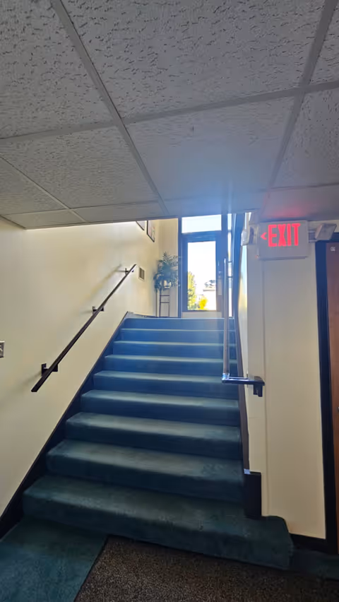 Blue-carpeted stairwell with handrails, ceiling tiles, and an exit sign at the top.