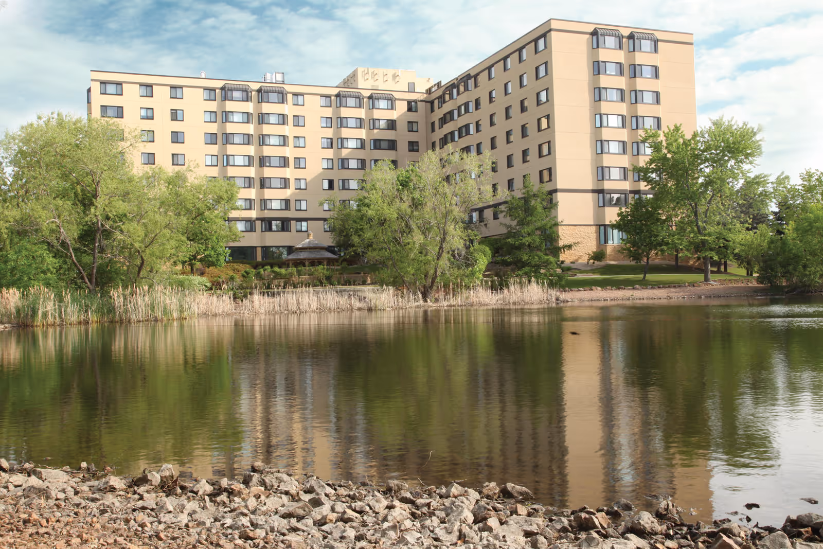 Exterior view of Ackerberg Family Sholom West Campus, a tan multi-story building beside a calm pond.