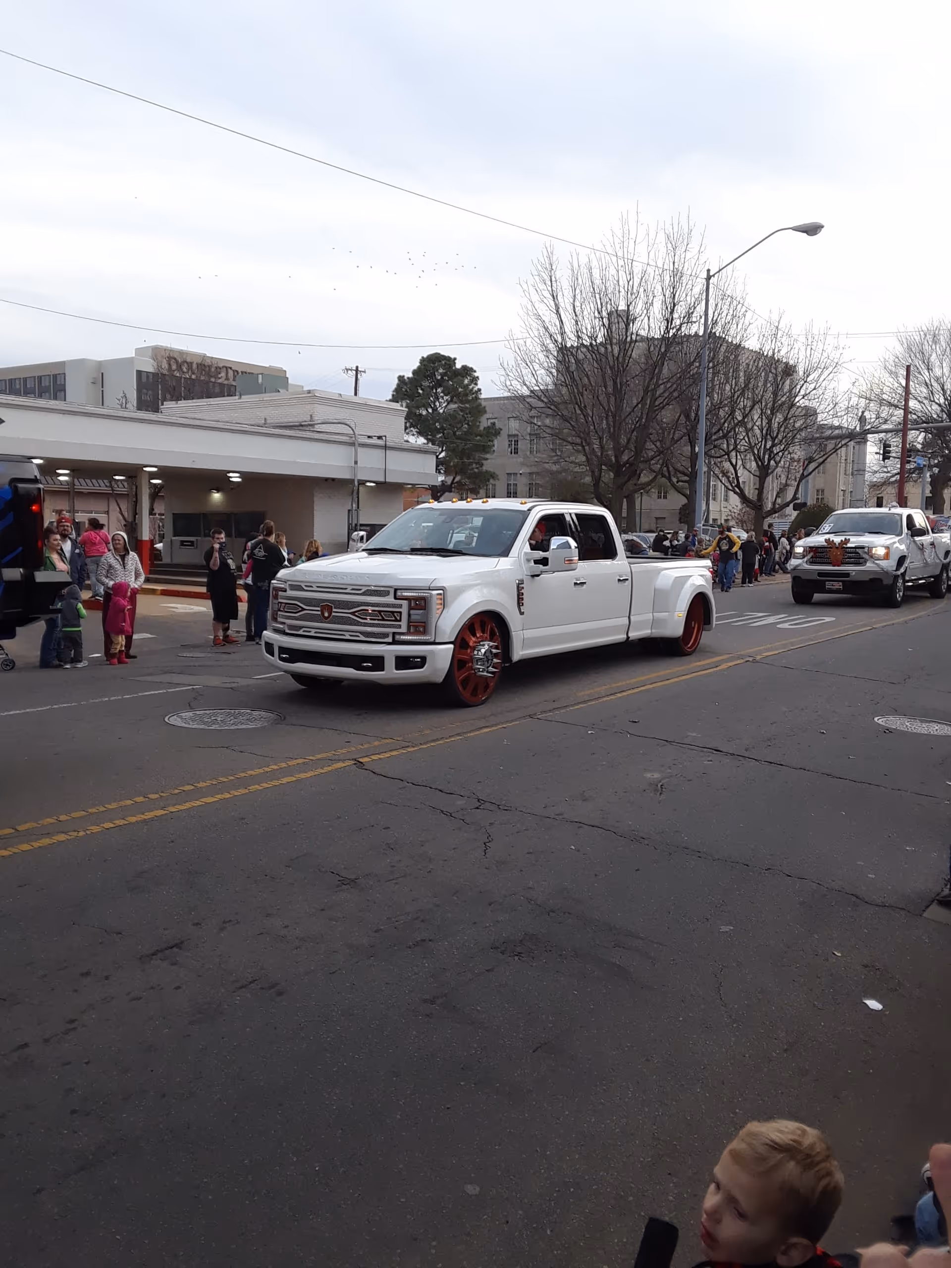 White pickup truck leads a parade down a city street with spectators and buildings.