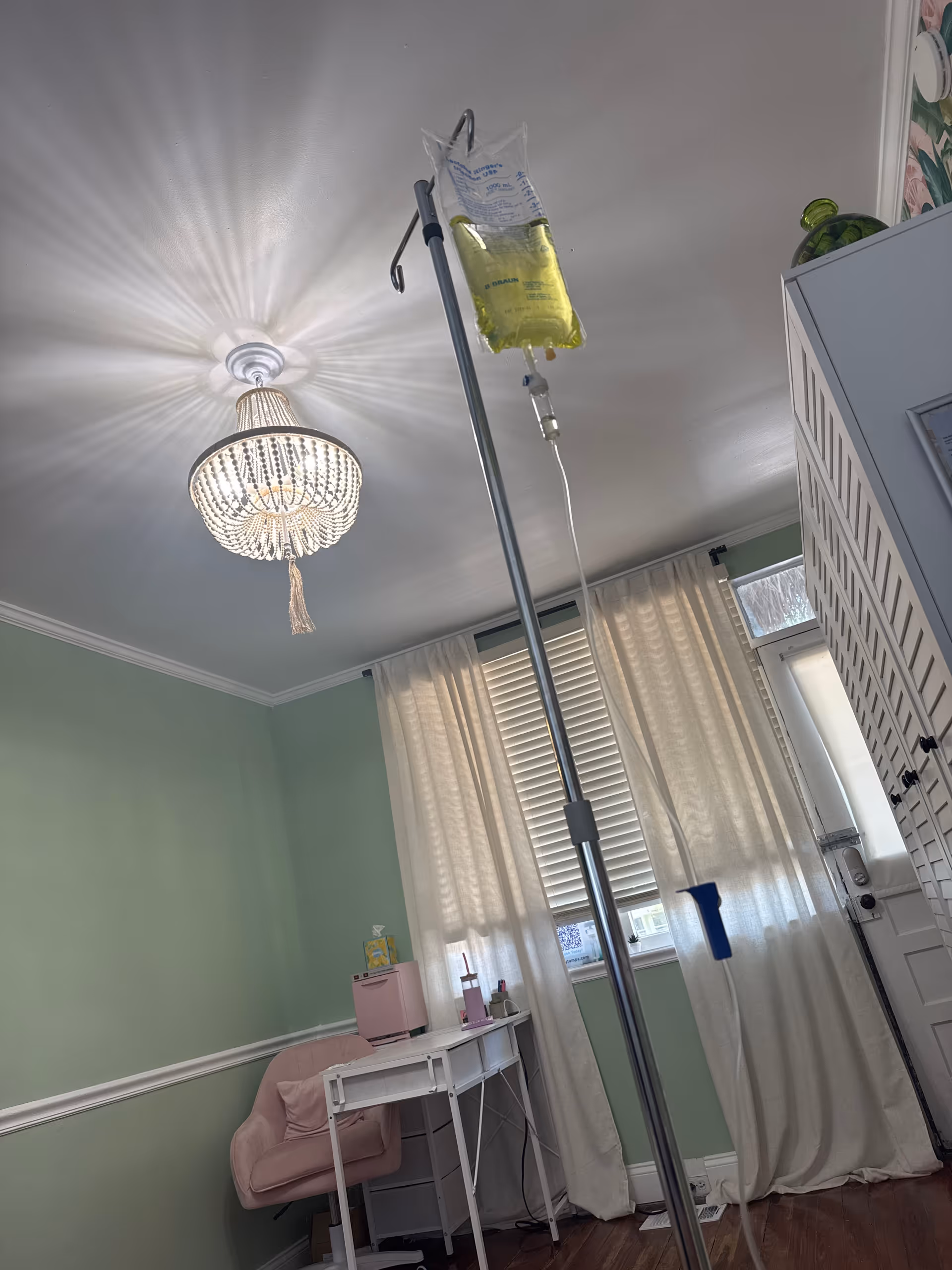 Pastel green patient room featuring an IV pole, chandelier lighting, desk, and curtained window.
