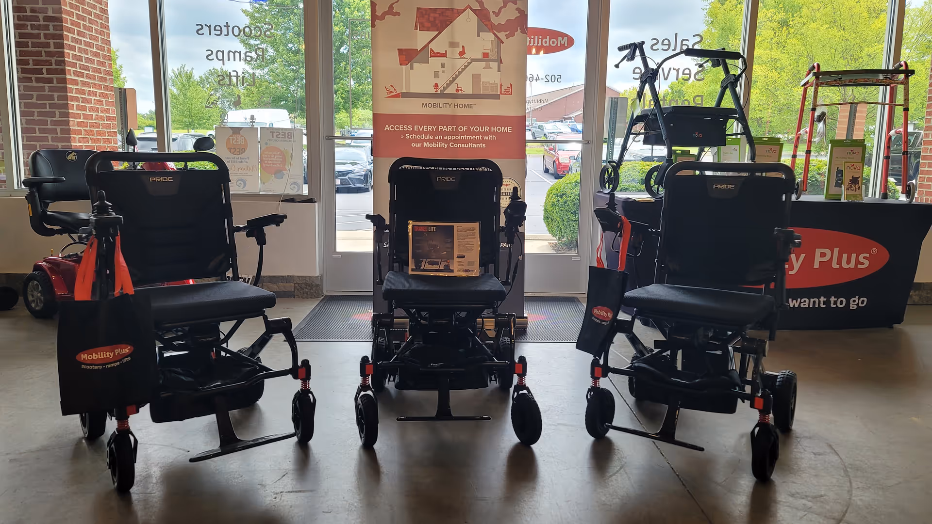 Three mobility scooters are displayed inside a Mobility Plus showroom near large windows.