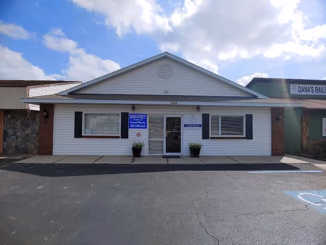 Single-story white building with blue shutters and central entrance, parking lot in front.