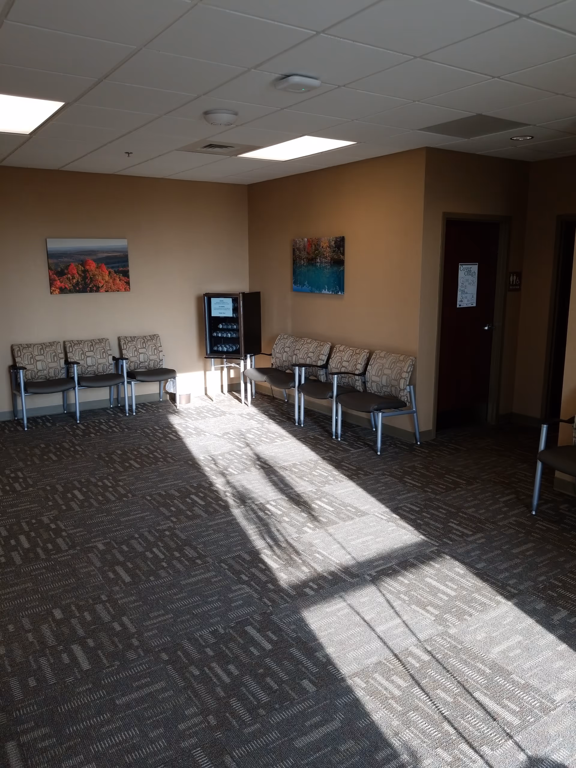 Empty waiting area in CoxHealth at Home with chairs, a beverage cooler, wall art, sunlight shadows.
