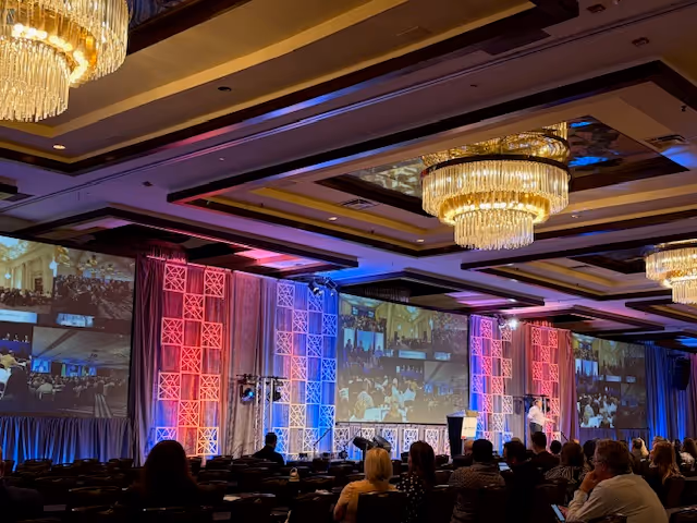 A large conference hall interior with chandeliers, a stage, and an audience.