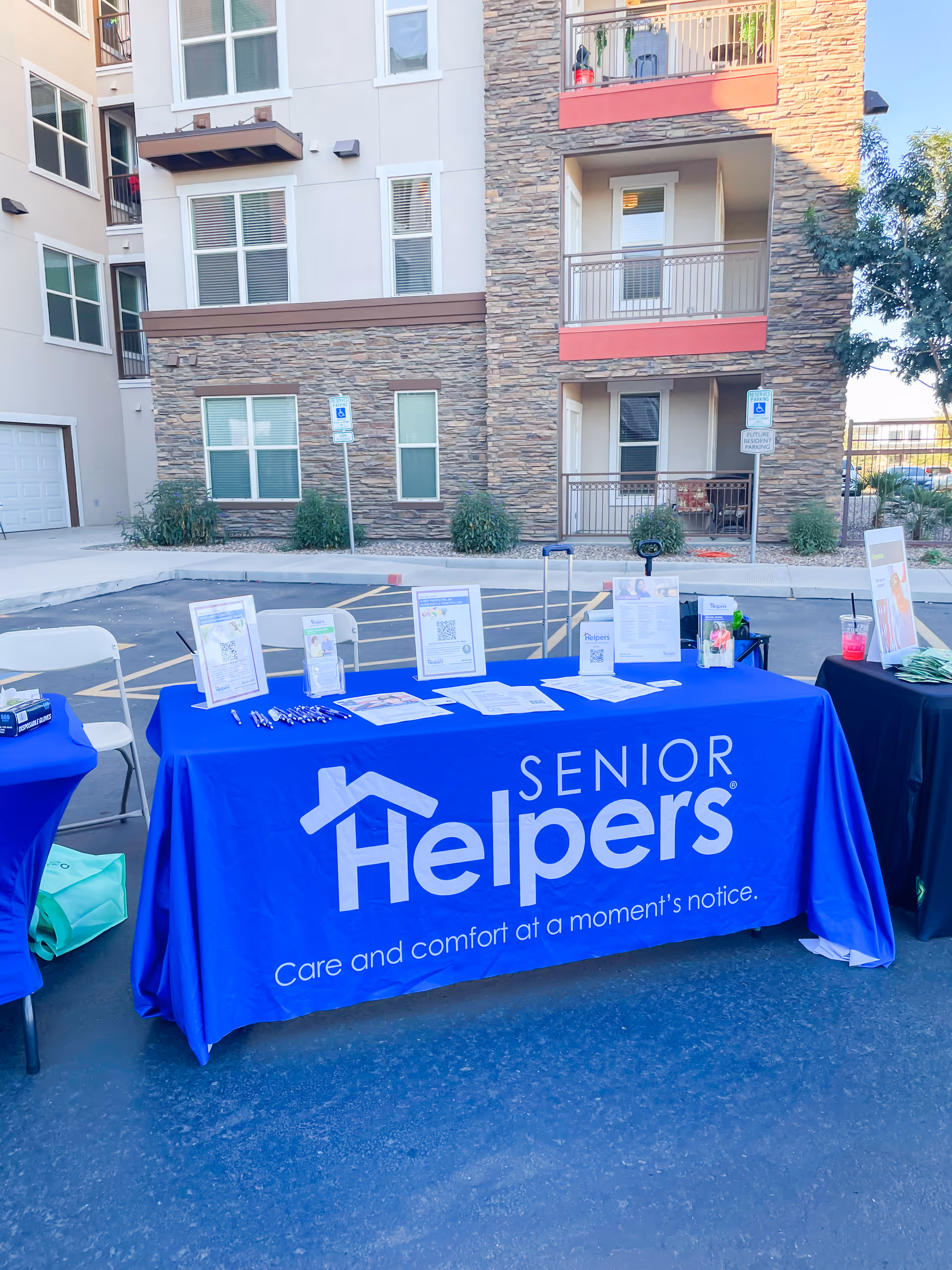 Outdoor promotional table with a blue Senior Helpers banner in a parking lot.
