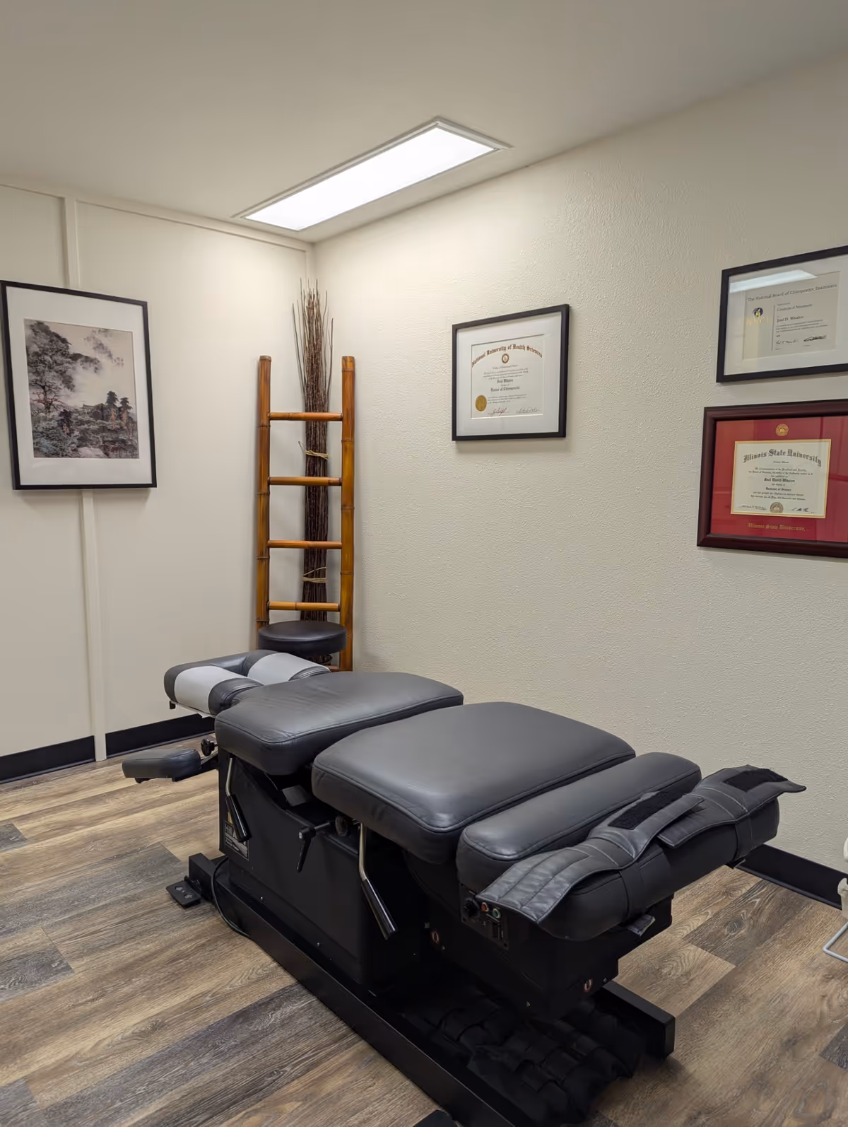 A chiropractic treatment table in a small exam room with framed certificates.