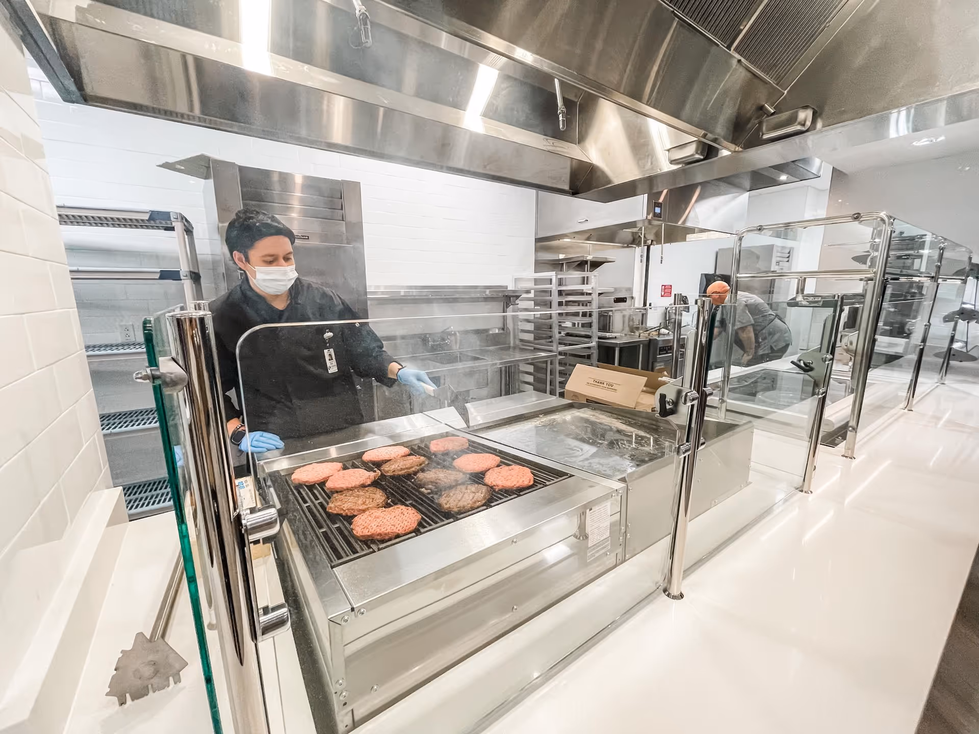 A chef wearing a mask grills burgers on a stainless steel kitchen station with a glass barrier.