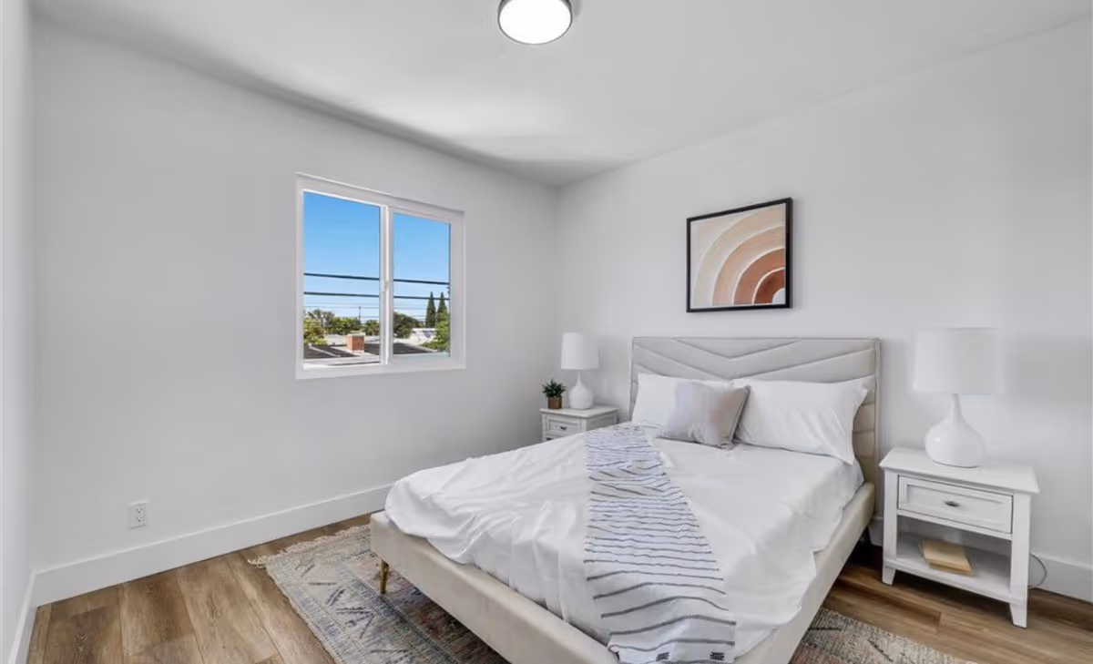 Bright white bedroom with a beige upholstered bed, nightstands, lamps, and abstract wall art.
