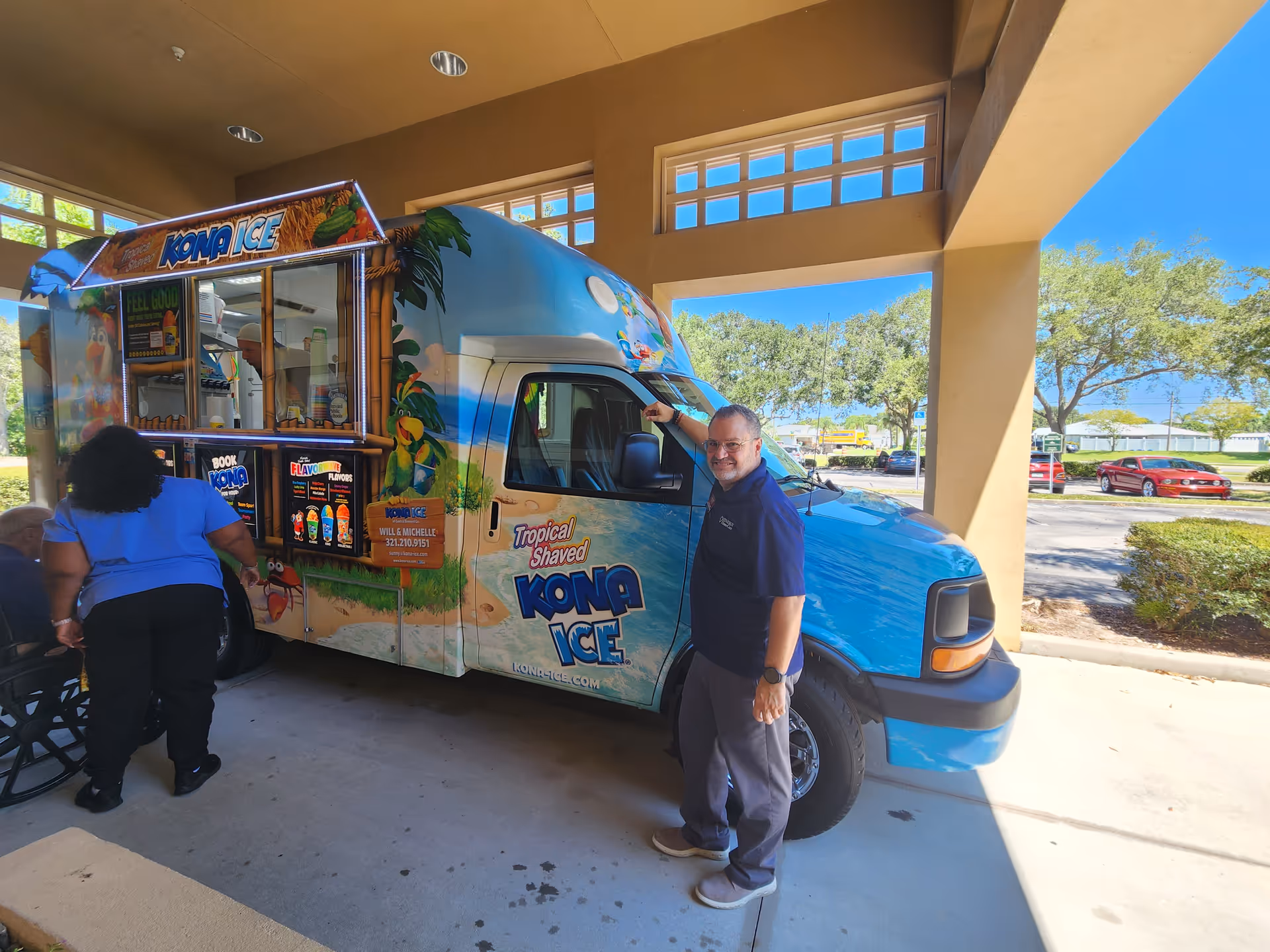 A colorful Kona Ice truck parked under a covered area with staff nearby.