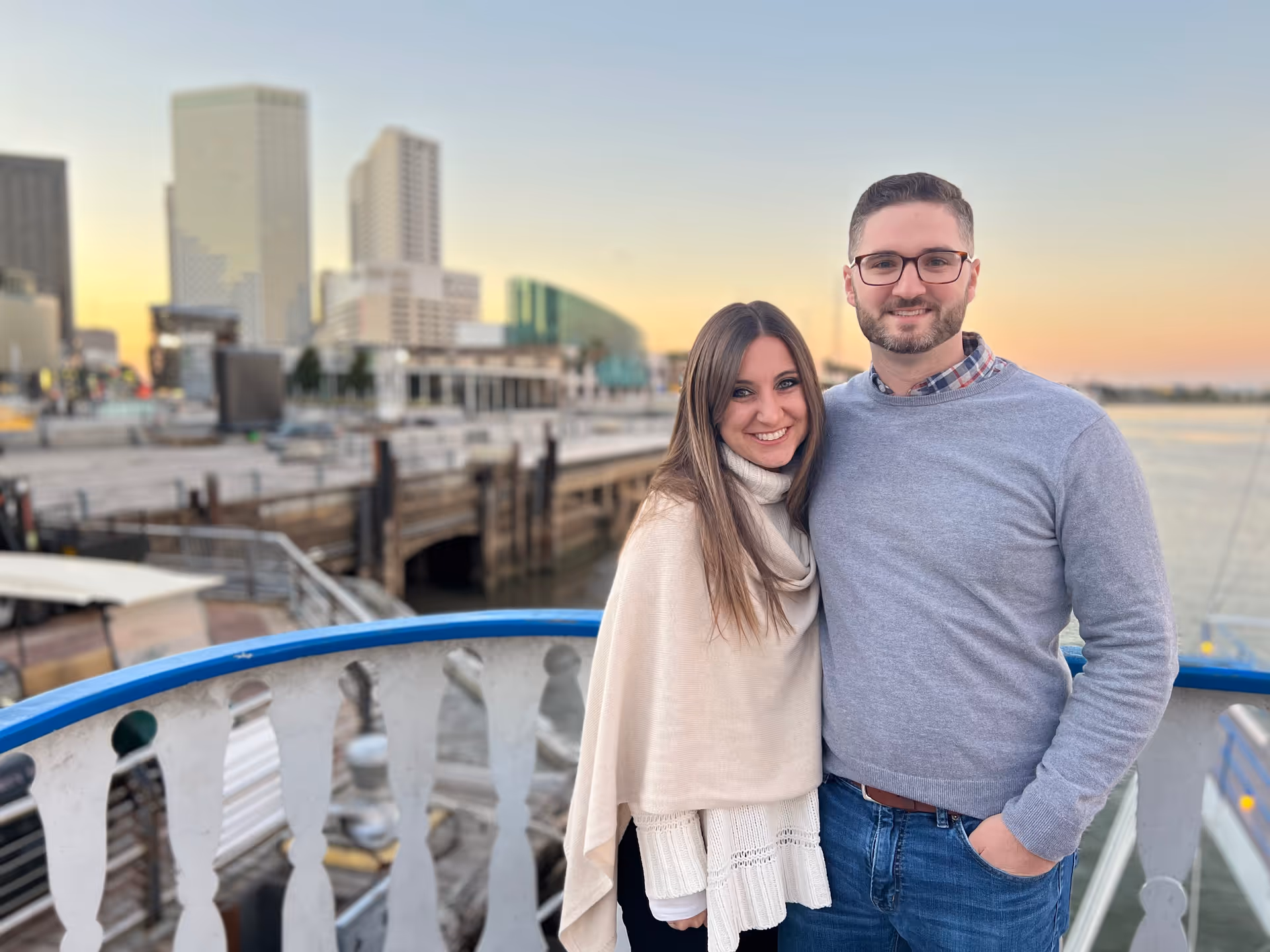 A couple stands on a waterfront balcony with a city skyline at sunset