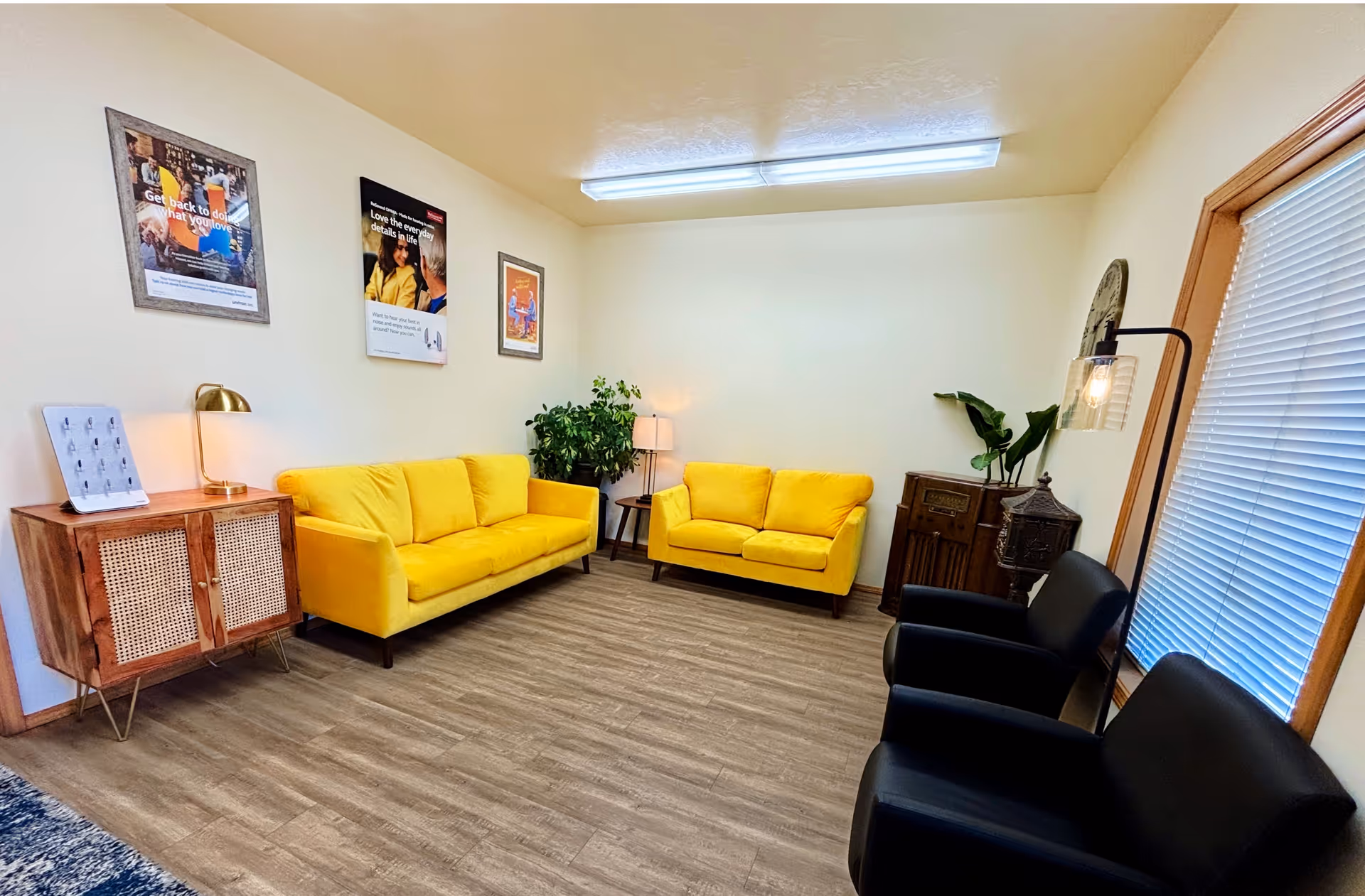 Interior waiting lounge with yellow sofas, black chairs, plants, and wall posters.