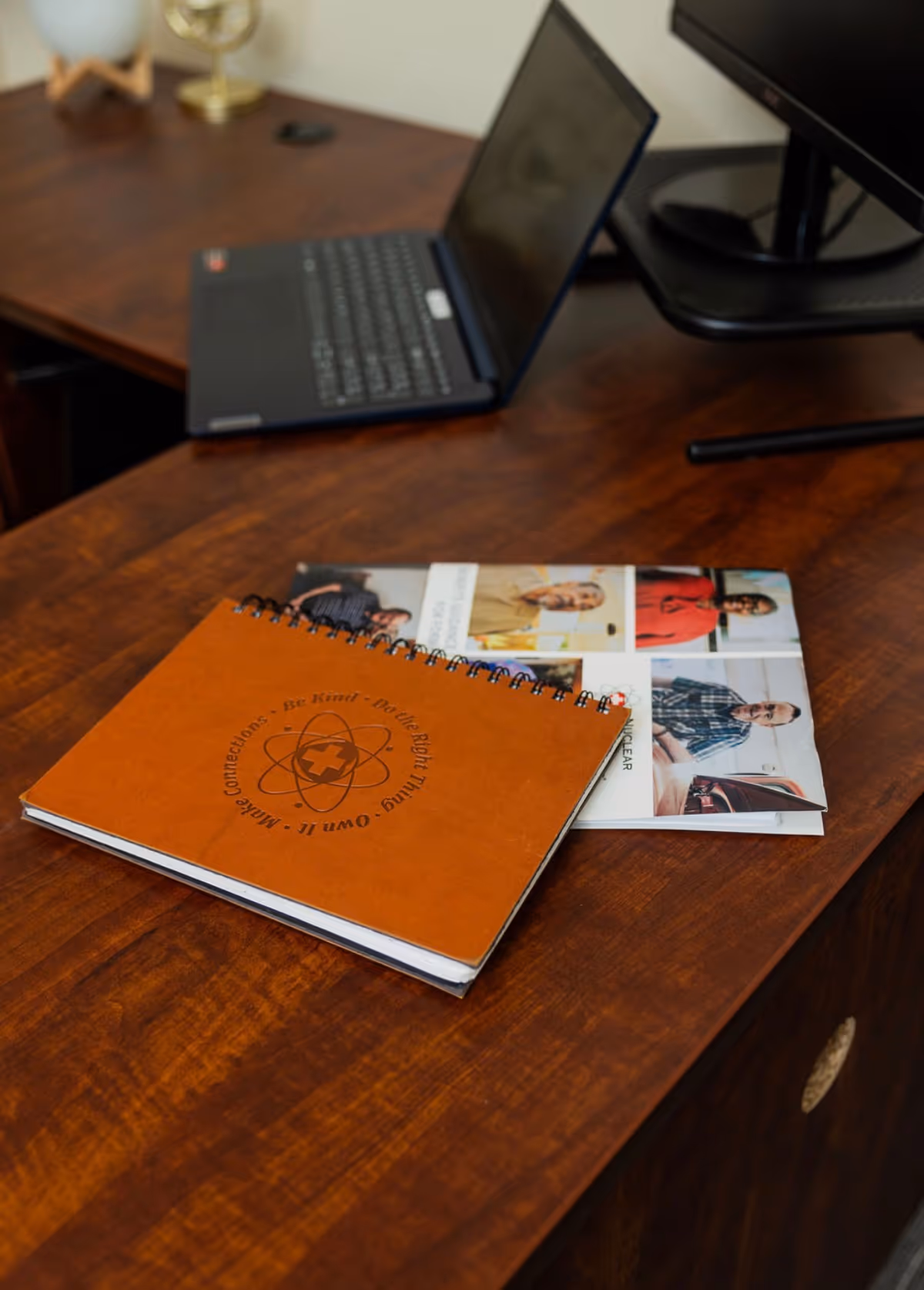 Wooden desk with laptop, monitor, and orange spiral notebook in a tidy office