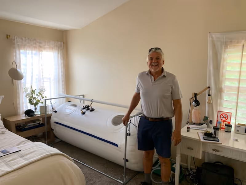 Smiling man stands beside a large white hyperbaric chamber in a cozy room