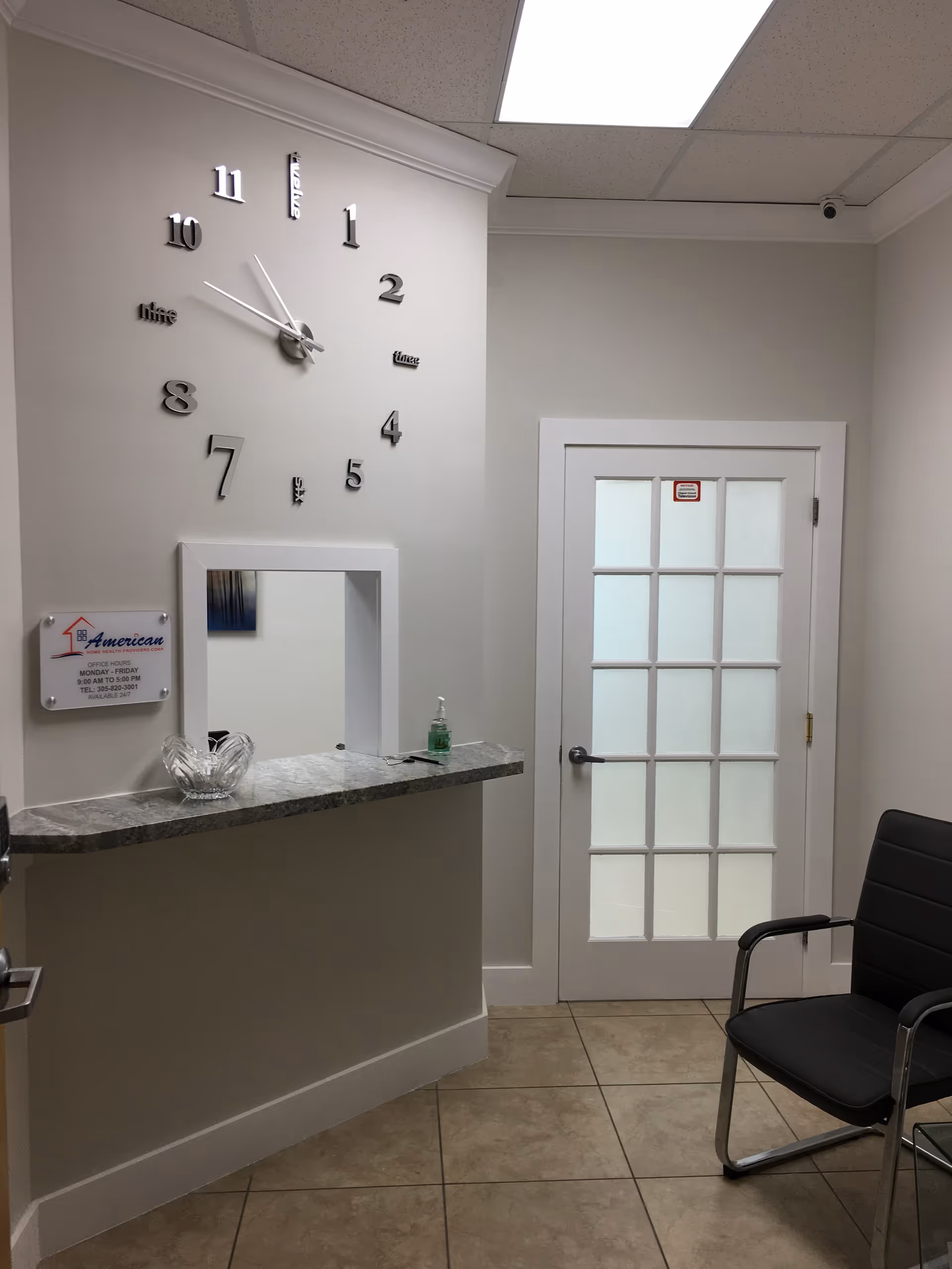 Reception area with a wall clock, marble countertop, glass-paneled door, and waiting chair.