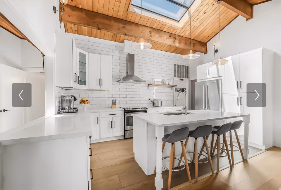 Bright white kitchen with island, stools, stainless appliances, and exposed wooden beams.