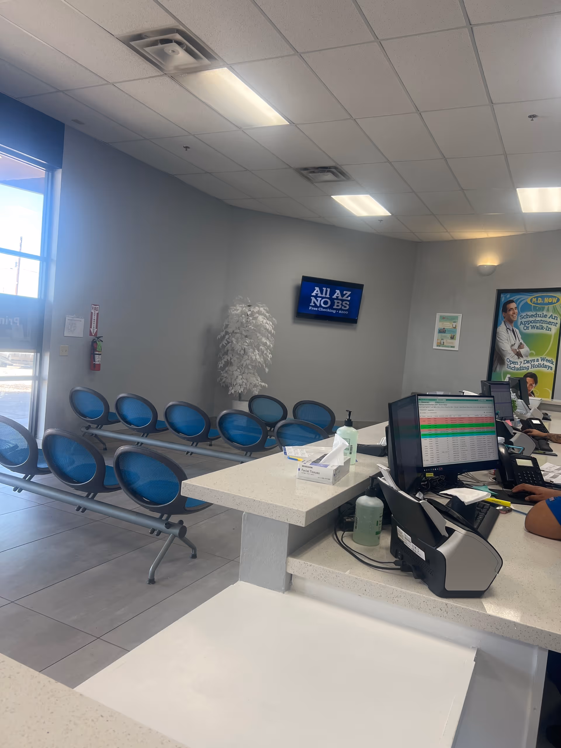 Reception area with computer, blue chairs, and a plant in a clinic lobby.