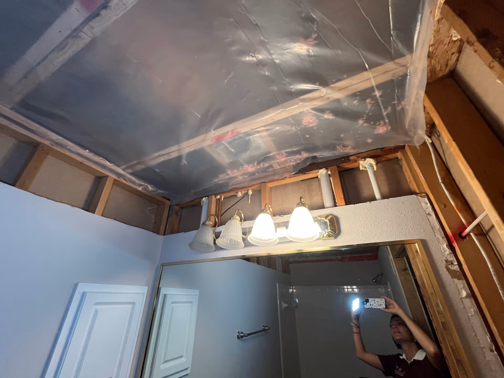 Bathroom under renovation with exposed framing, plastic ceiling, and a person taking a mirror selfie.