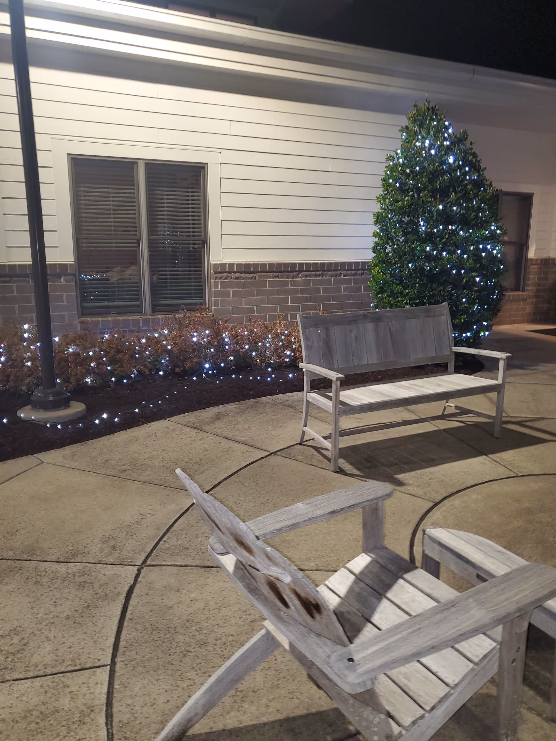 Exterior view of Alive Hospice Murfreesboro with lit shrubs, tree, and wooden benches.