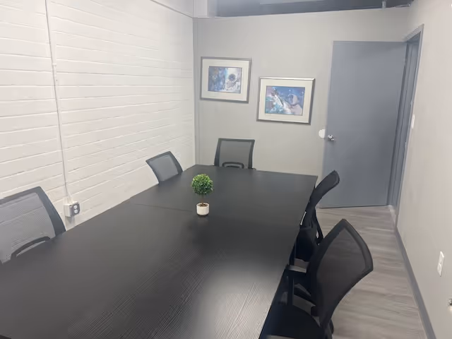 A long dark conference table with chairs and a plant is in a gray meeting room.